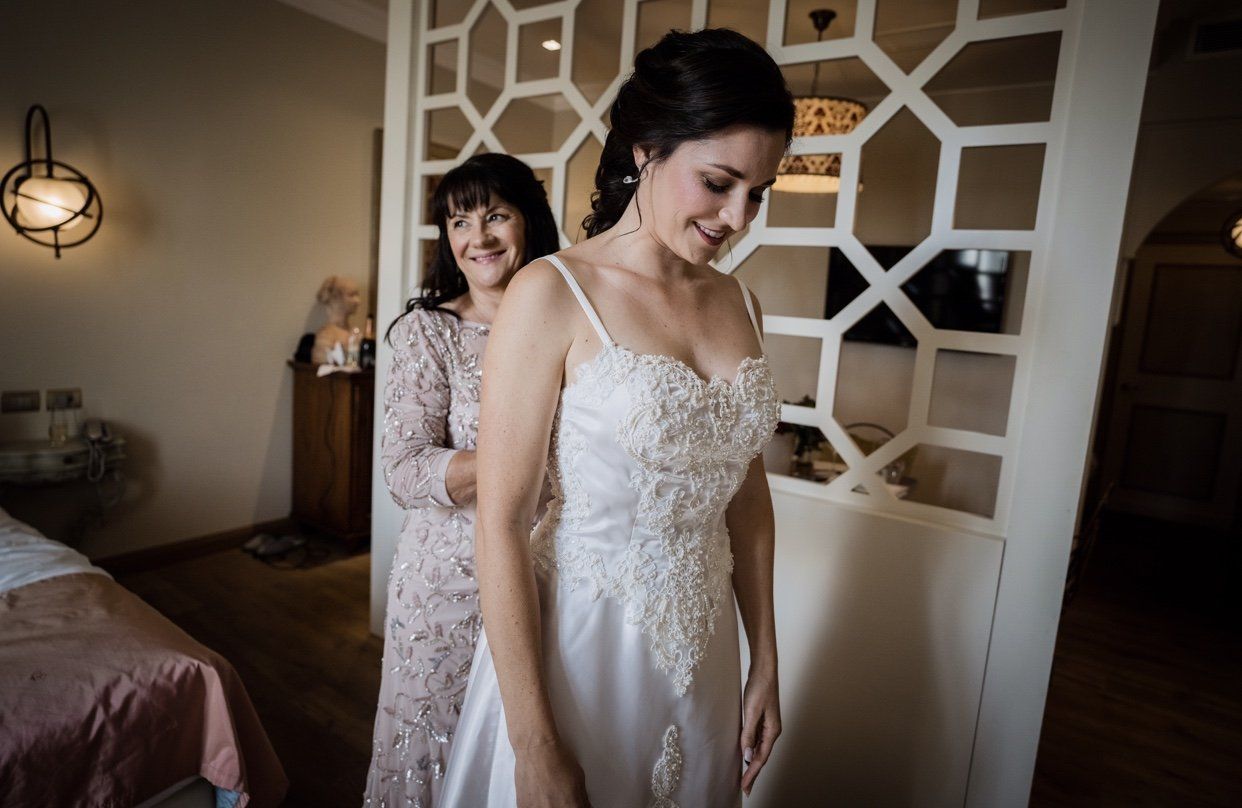 Bride in a white dress stands smiling while another woman helps her get ready in a bedroom