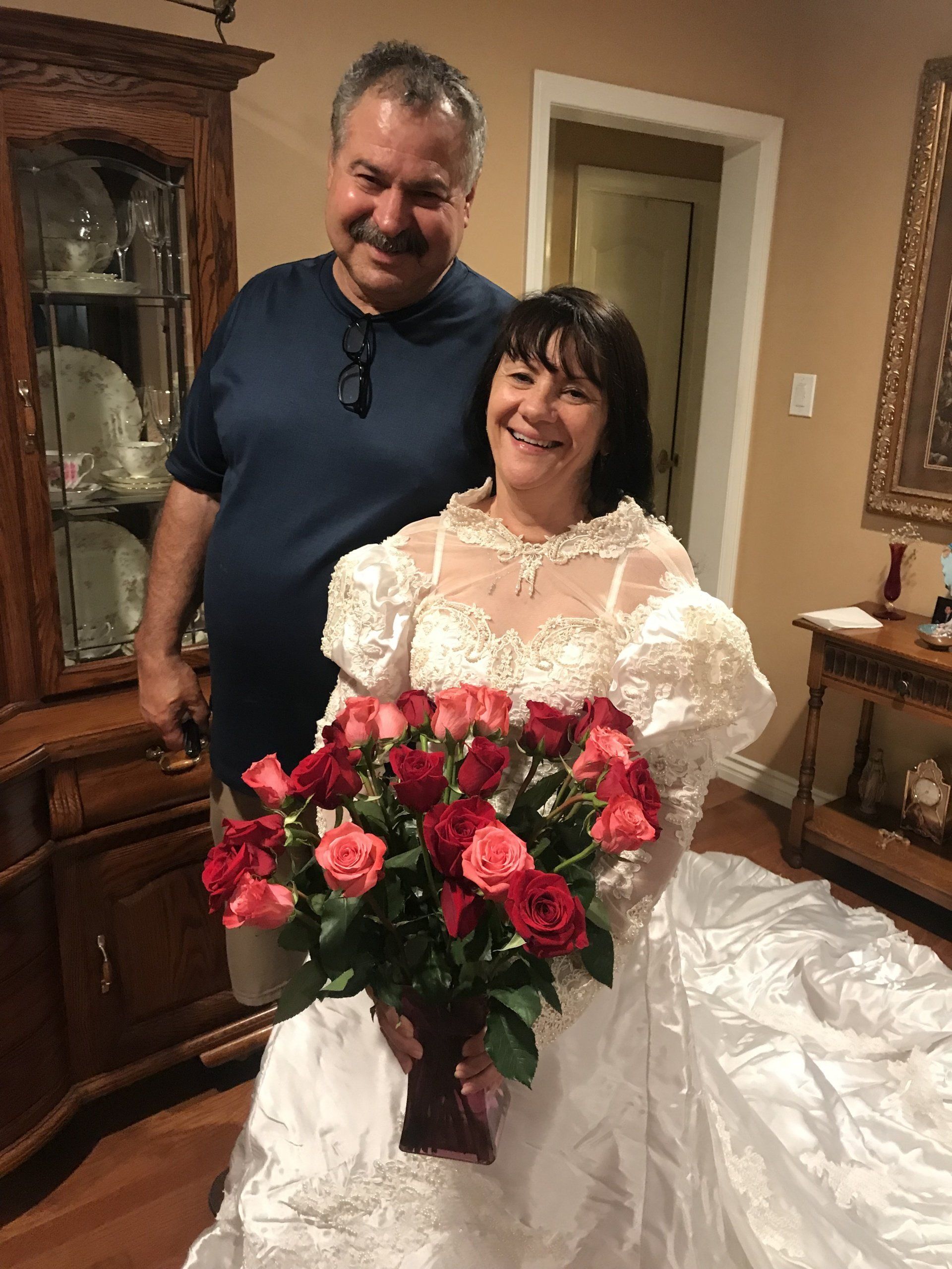 Bride in a white gown holding red roses, standing beside a smiling man indoors