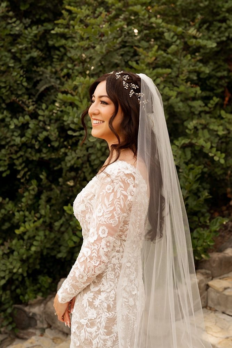 Bride in a white lace dress and veil standing outdoors, smiling in profile.