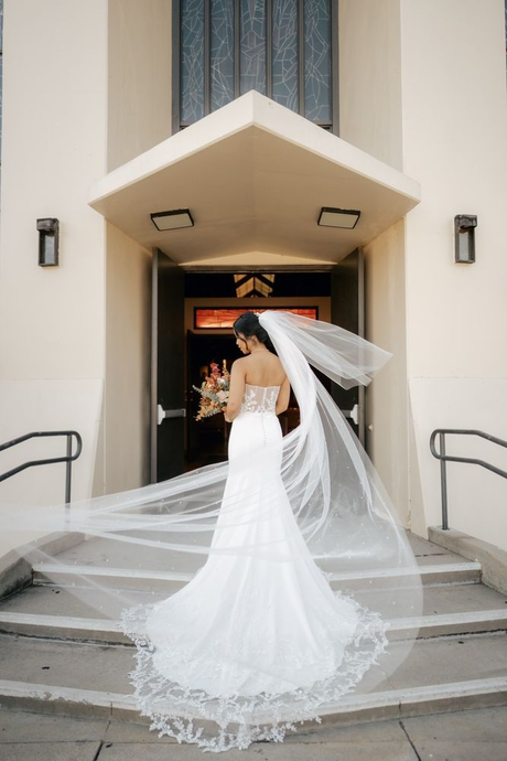Bride in white gown with long veil standing at church entrance, viewed from behind