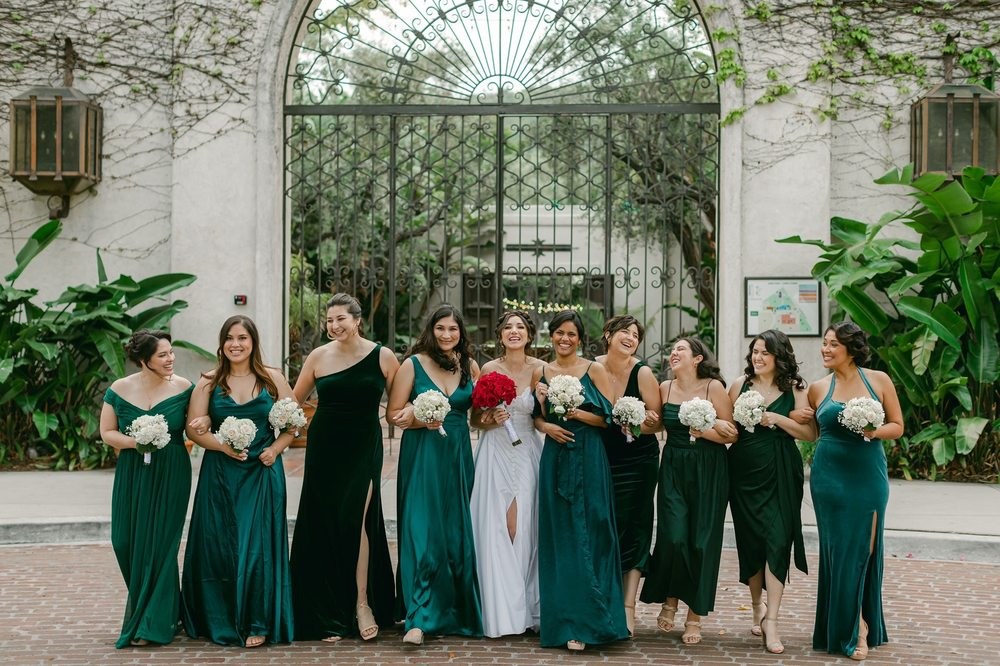 Bridal party in green dresses stands holding bouquets in a garden courtyard before an arched gate