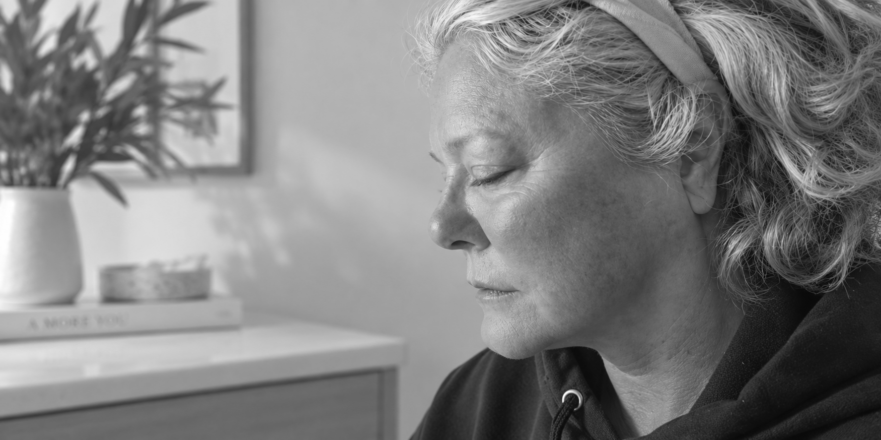 Woman in profile with eyes closed, seated beside a potted plant in a bright room during a cheek filler appointment.