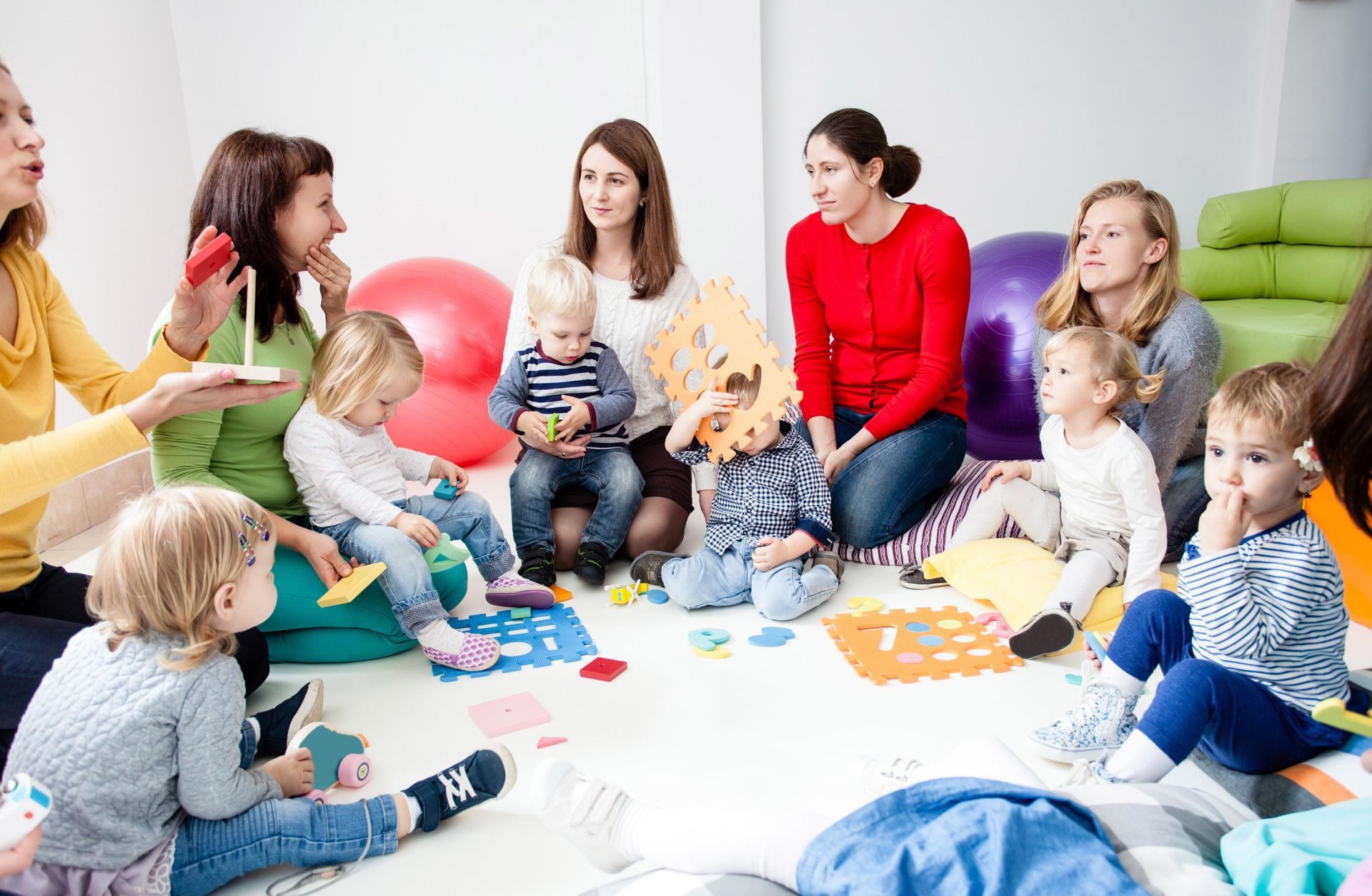 Group of mothers and toddlers in a playroom; women are seated around children, interacting and playing.