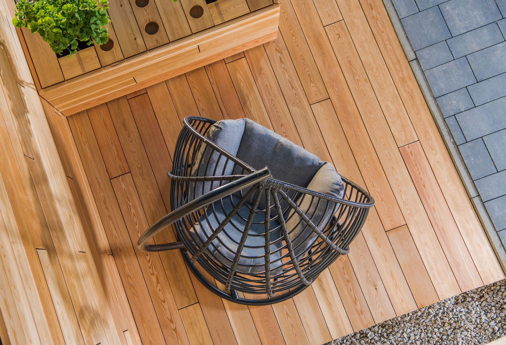 Top-down view of a woven armchair with cushions on a wooden deck next to a planter and tiled path.