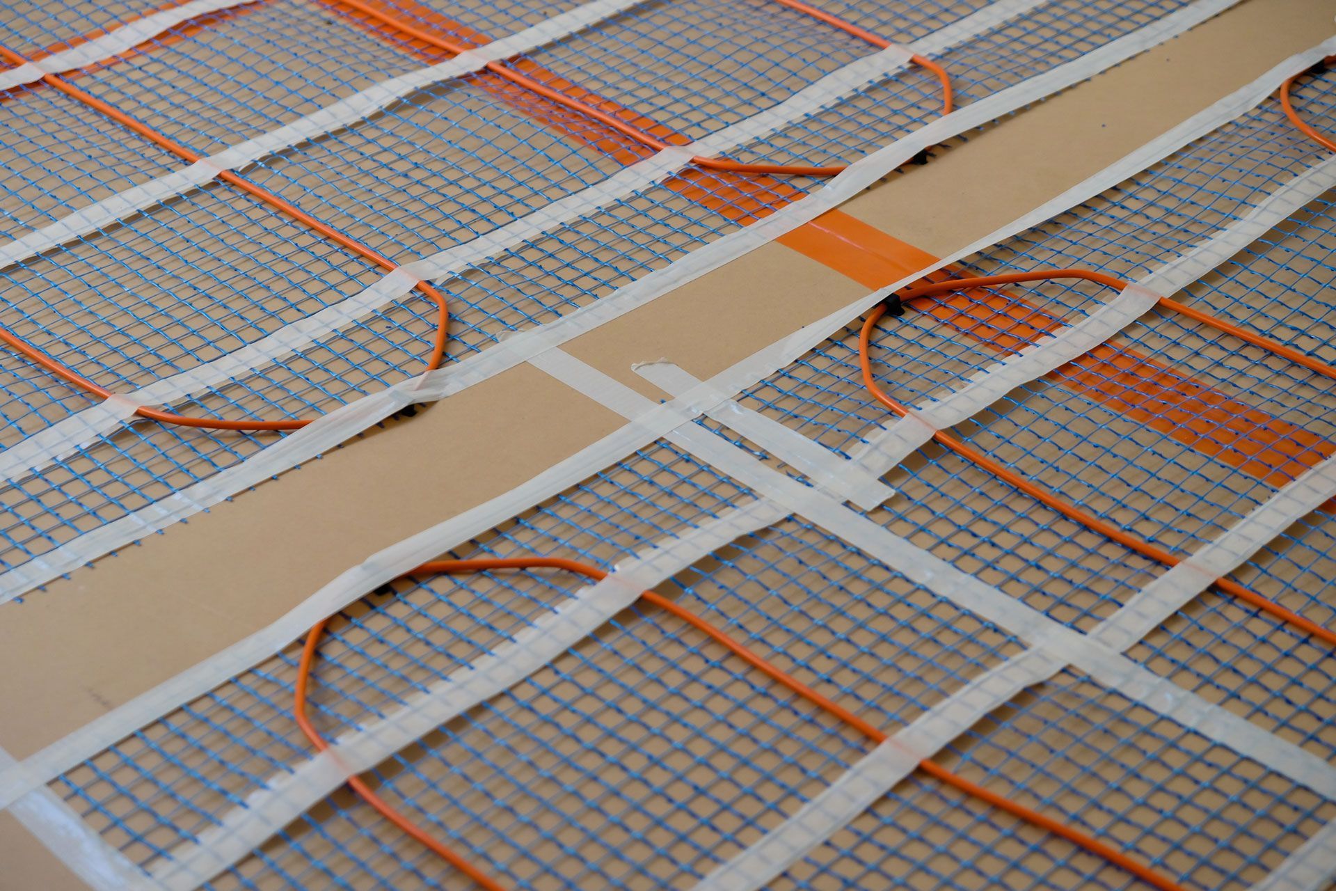Electric floor heating system: orange wires on a mesh mat, secured with white tape.