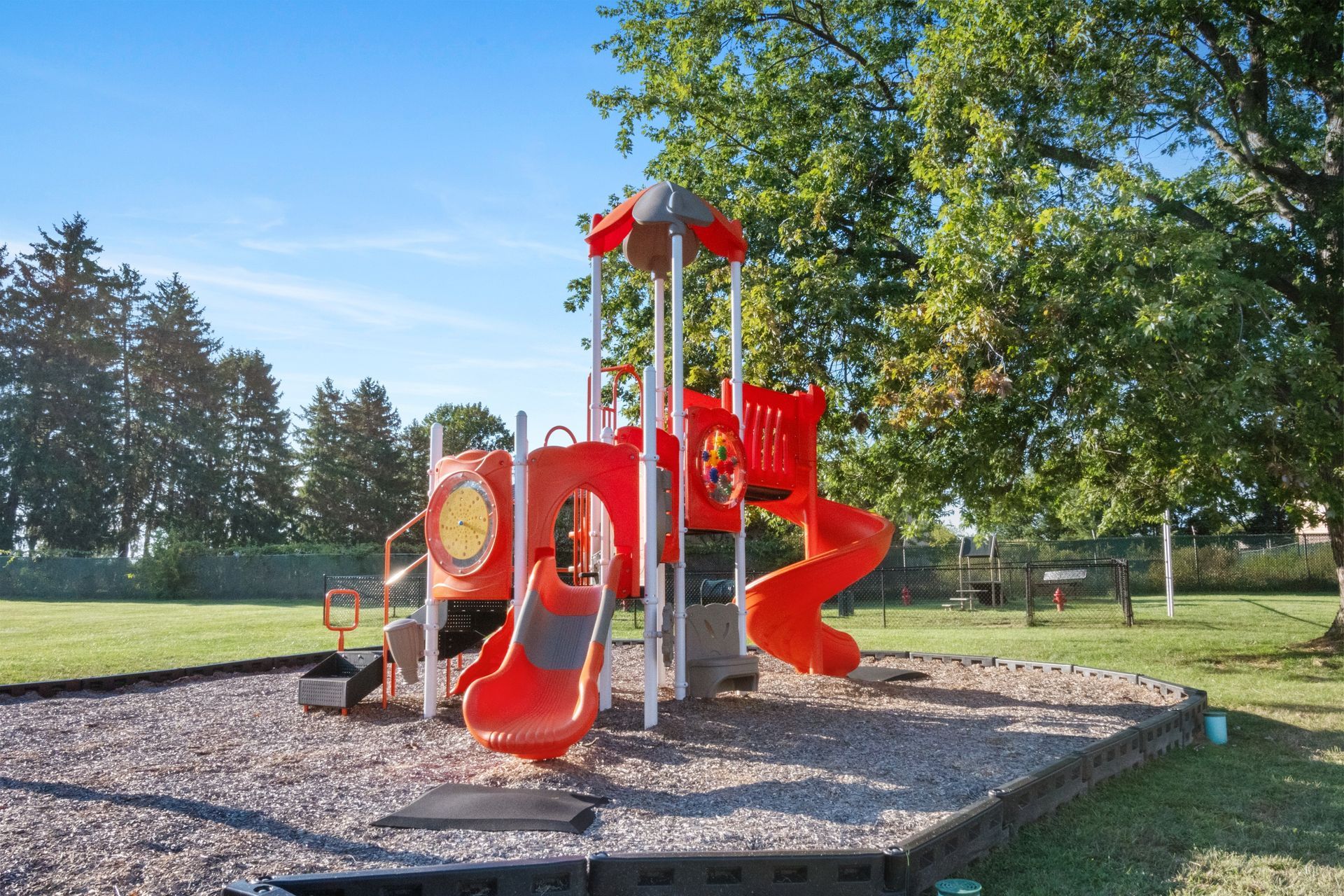 Bright red playground slide and climbing structure in a sunny park with trees and wood chips