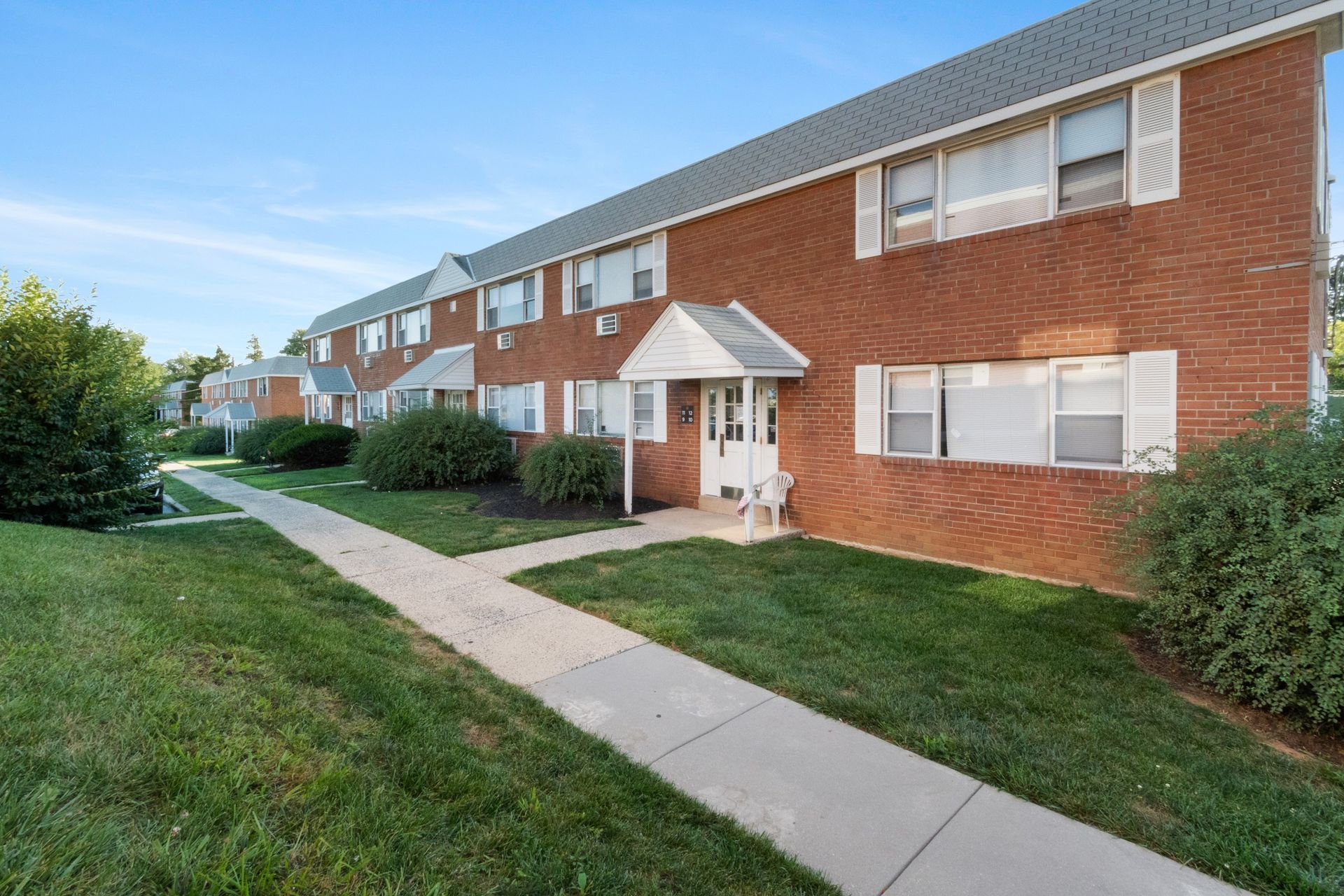 Brick apartment building with white trim, front walkway, and green lawns under a clear blue sky