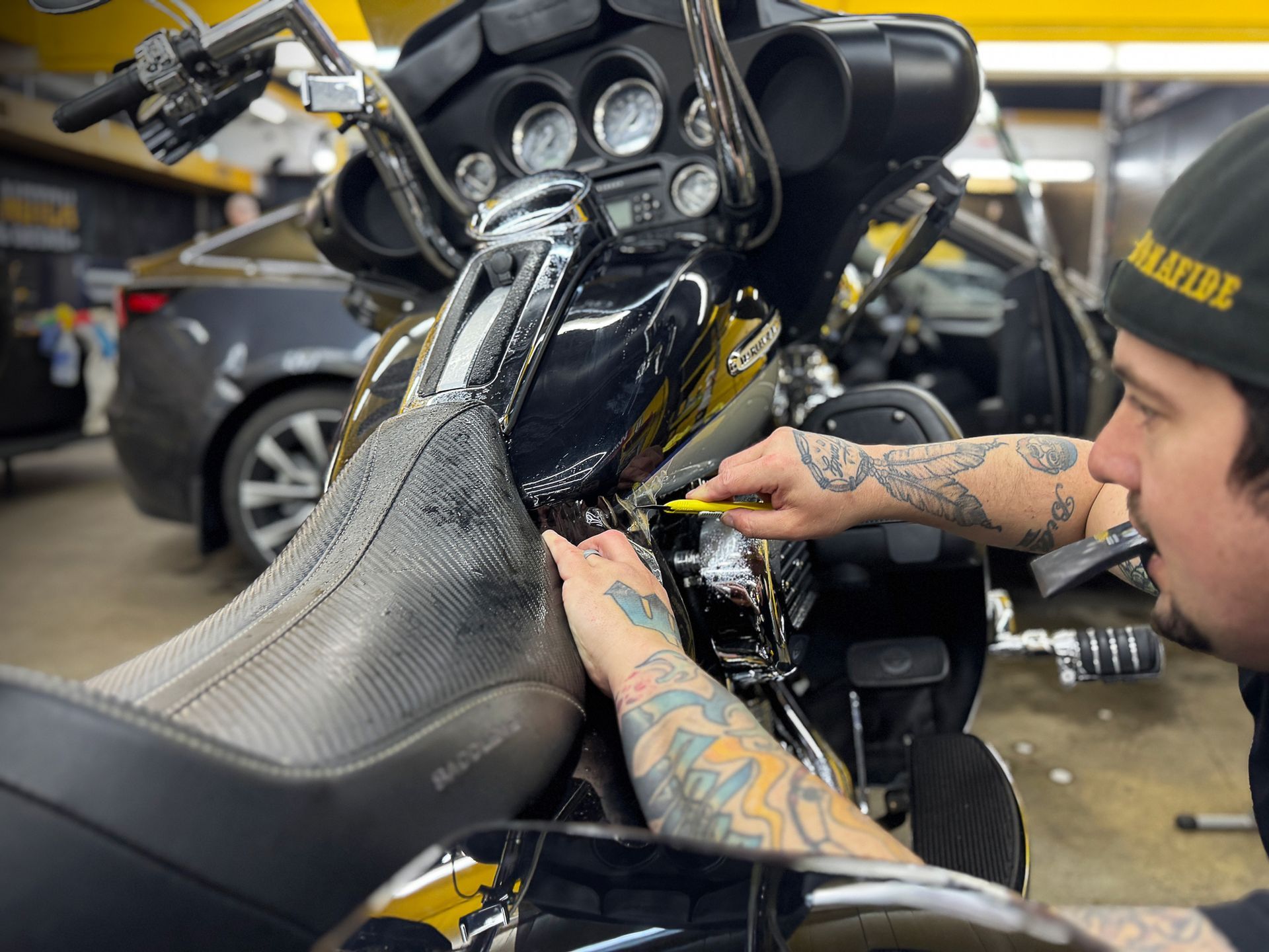 Technician polishing a black motorcycle fairing in a garage, with tattooed hands visible.