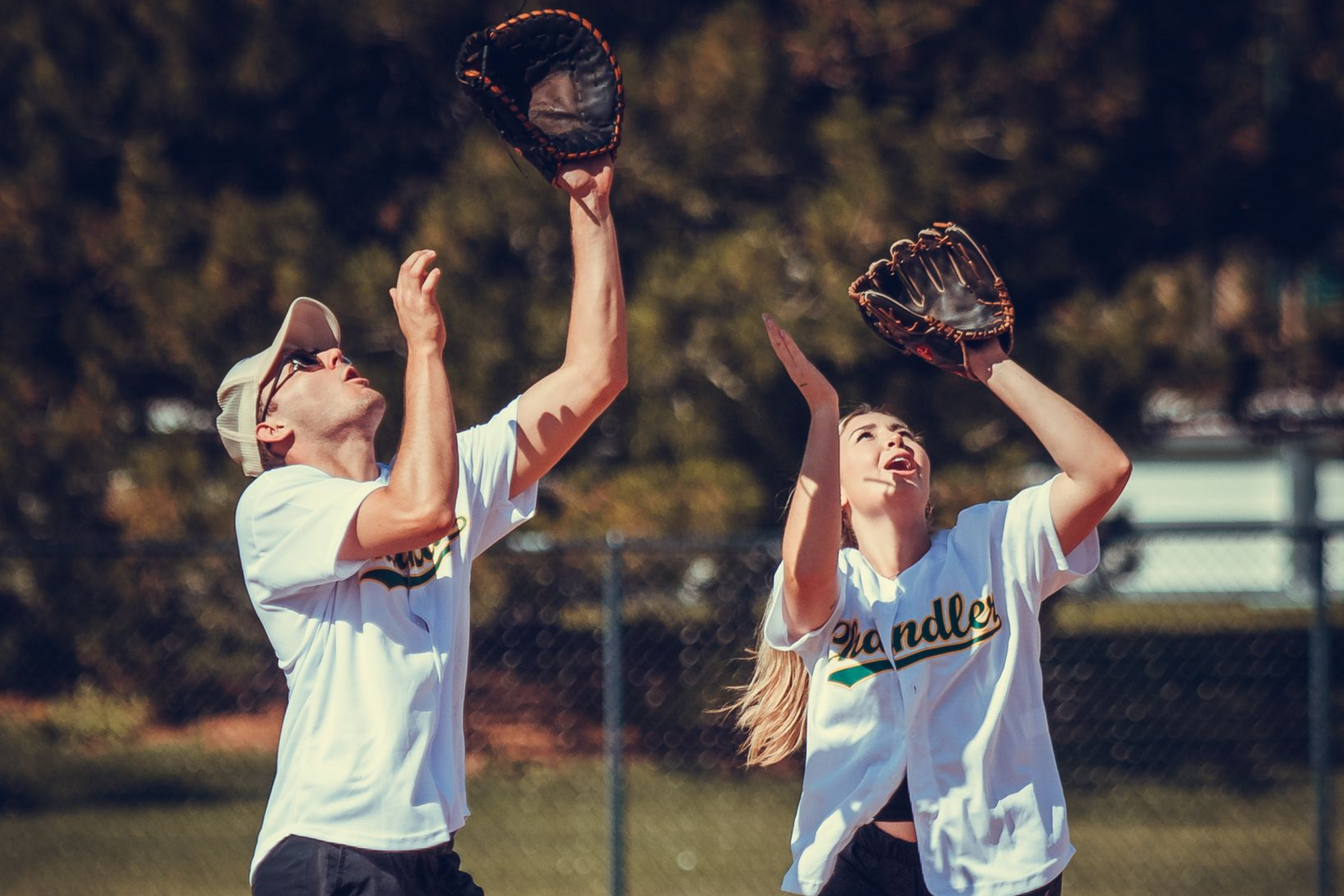 Two softball players in white jerseys catch a ball with gloves raised on a sunny field