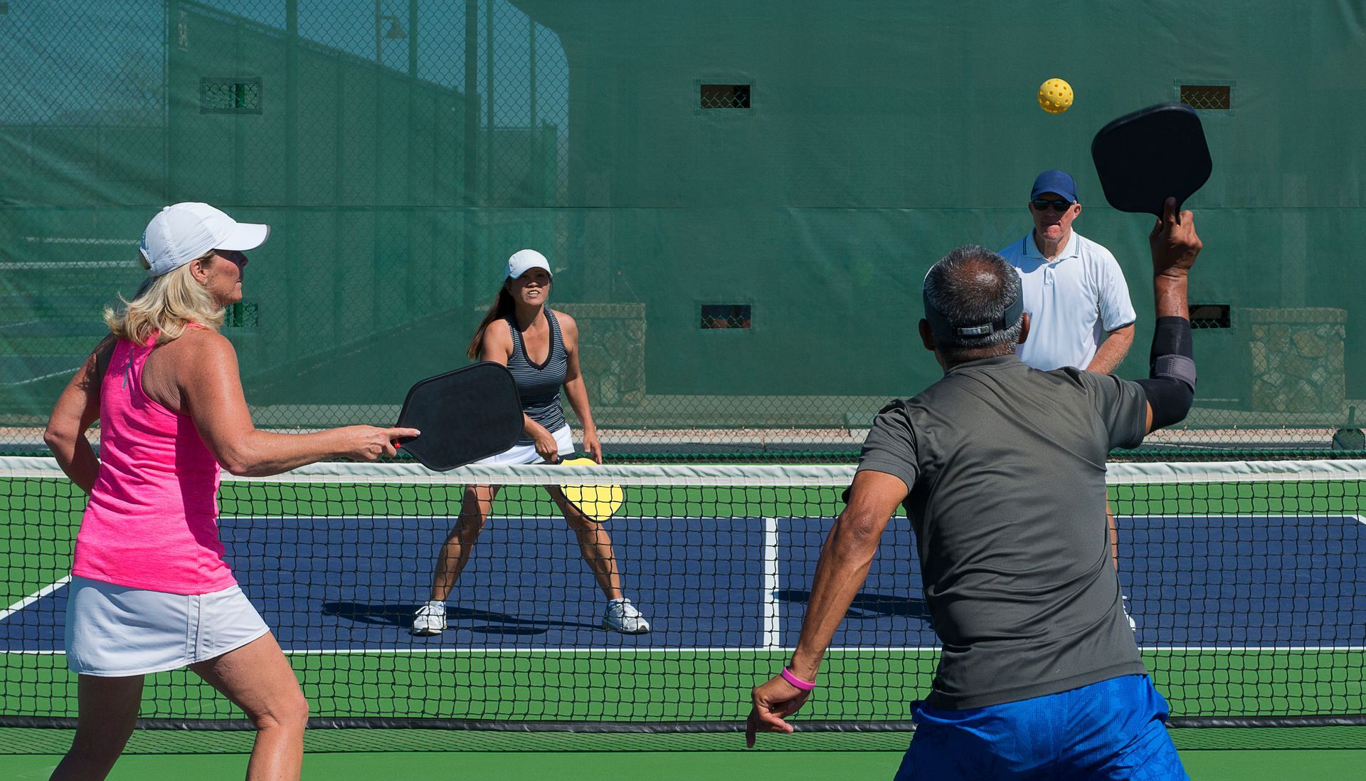 Four people play tennis on an outdoor court, with one player hitting a yellow ball near the net.