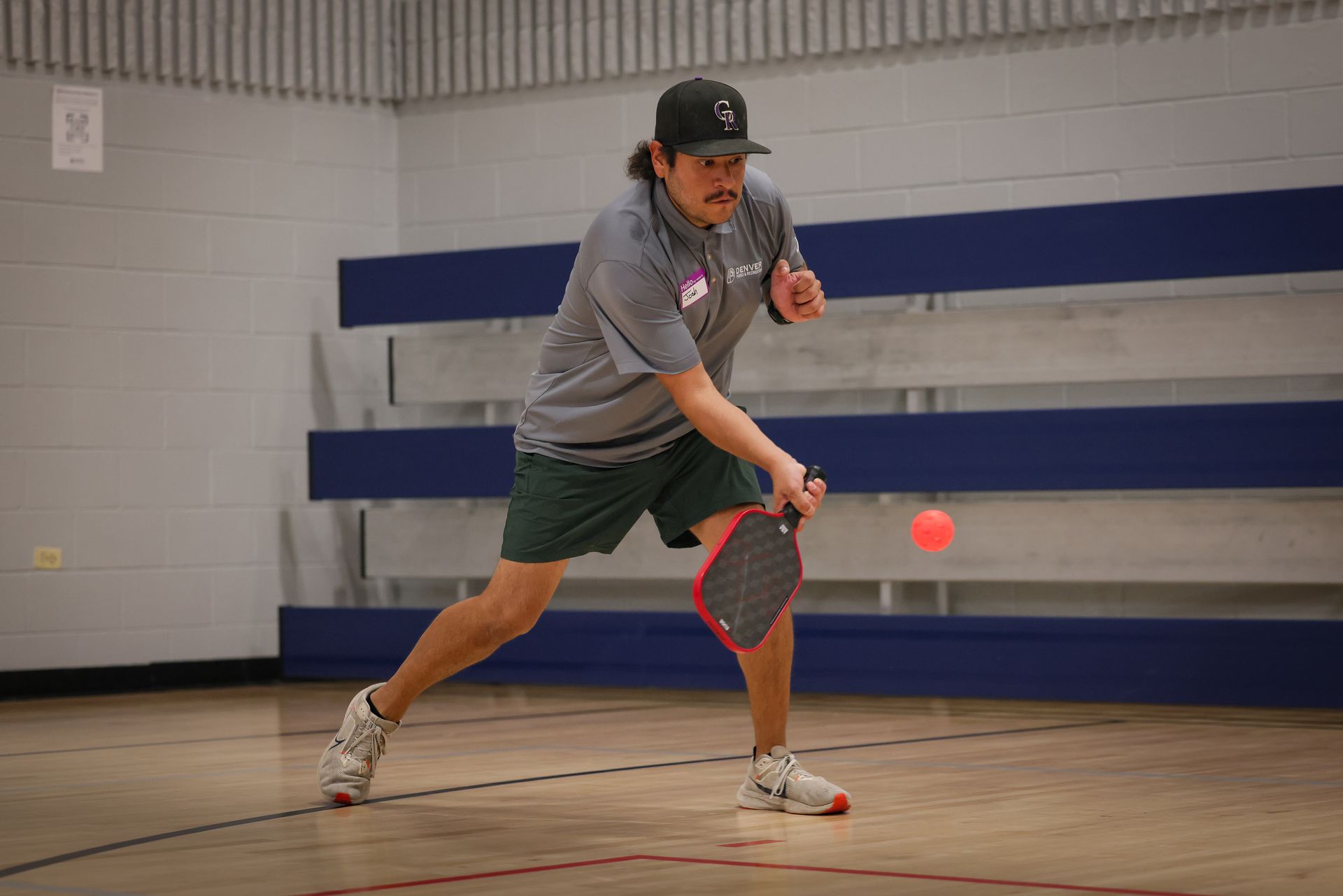 Person in gray shirt lunging to hit a pickleball in an indoor gym with blue wall padding.