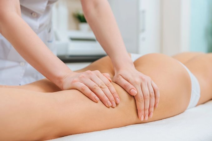Un'estetista applica uno scrub esfoliante sulla coscia di una persona sdraiata su un lettino bianco.