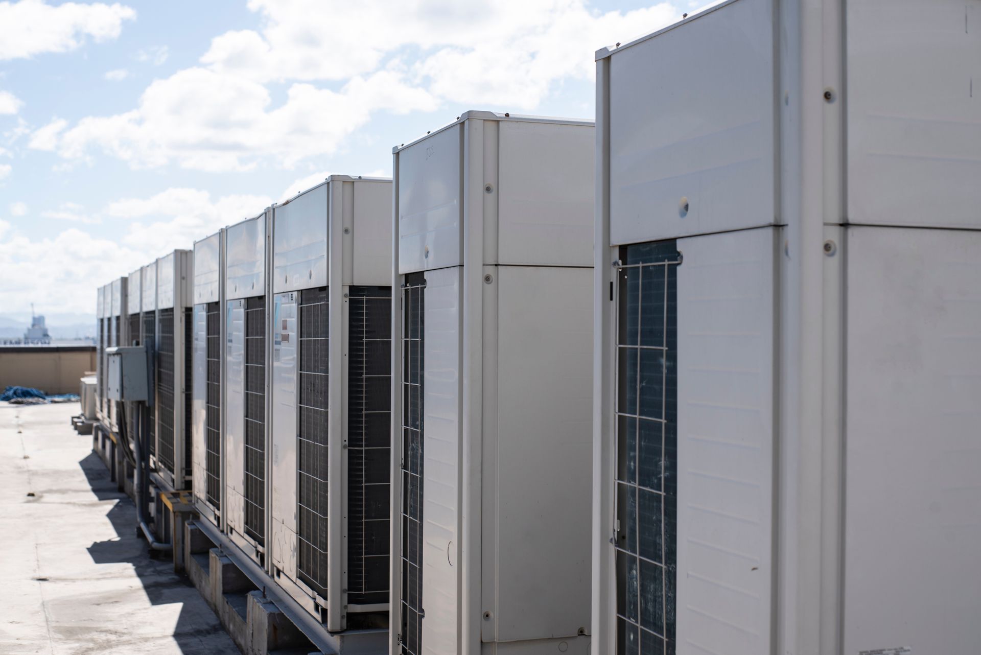 Row of large rooftop HVAC units beside a building under a partly cloudy sky Row of large rooftop HVAC units beside a building under a partly cloudy sky