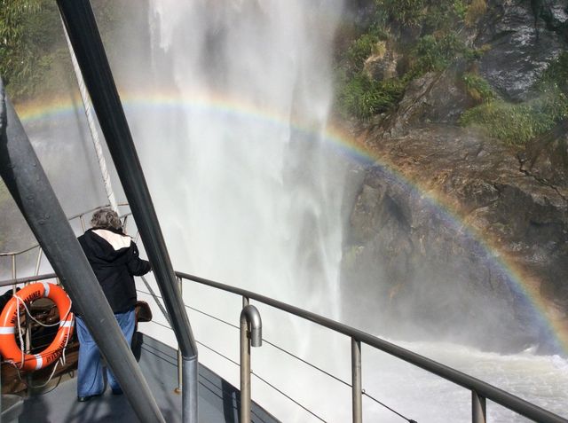 A passenger on a cruise boat deck beside a waterfall and a rainbow