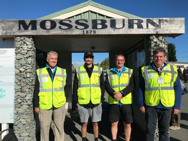Four tour guides in high-visibility vests under a sign reading Mossburn 1879