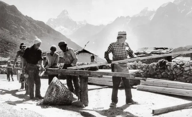Black-and-white photograph of volunteers cutting timber planks at the Khunde Hospital building site, mountains behind