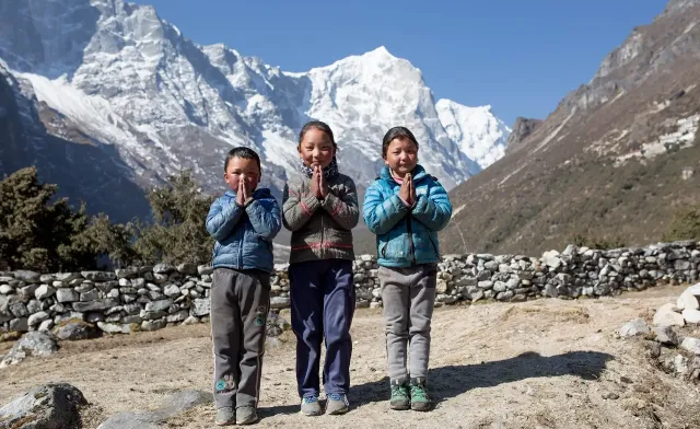 Three schoolchildren with their hands together in greeting, snow-capped peaks behind