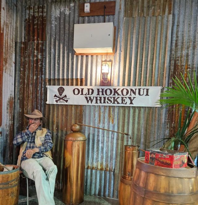 A seated figure beside whiskey barrels under an Old Hokonui Whiskey sign