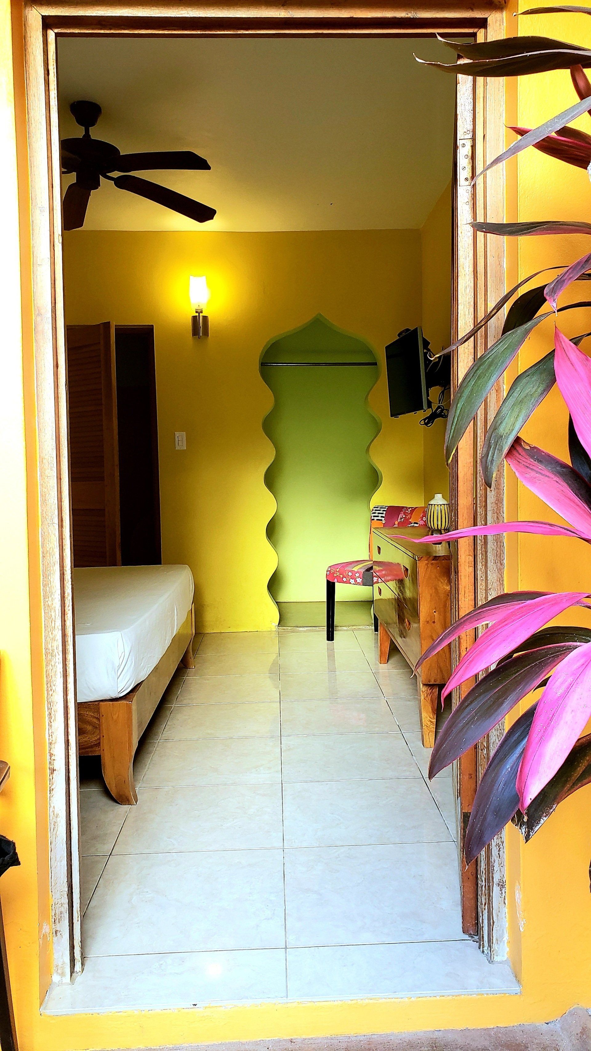 View of a brightly lit room through an open doorway, yellow walls and tiled floor, with a bed and table visible.