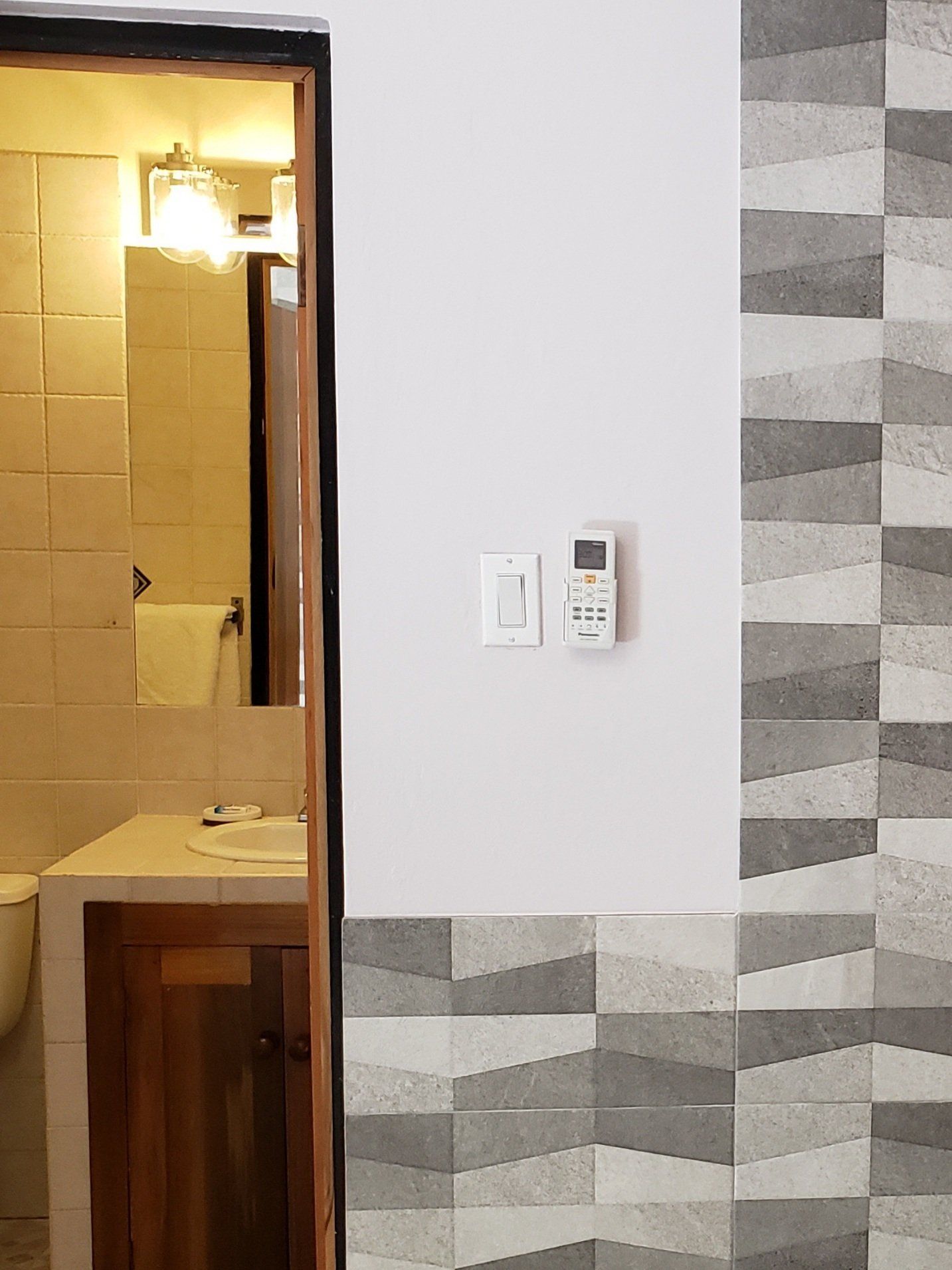 Bathroom doorway, white and patterned gray walls, light switch, AC remote, wooden cabinet.