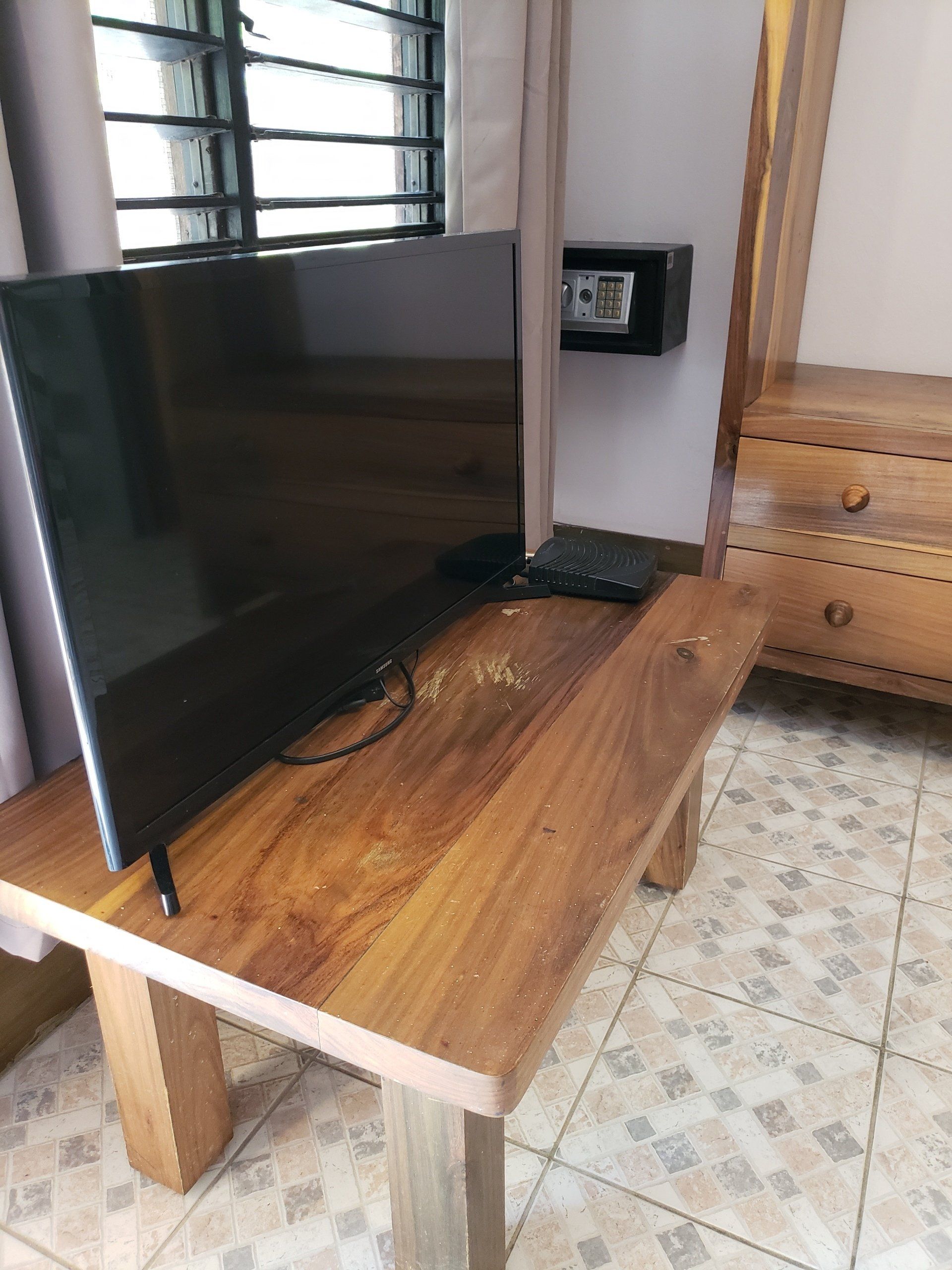 TV on a wooden table, window in the background, safe on wall, dresser to the side.