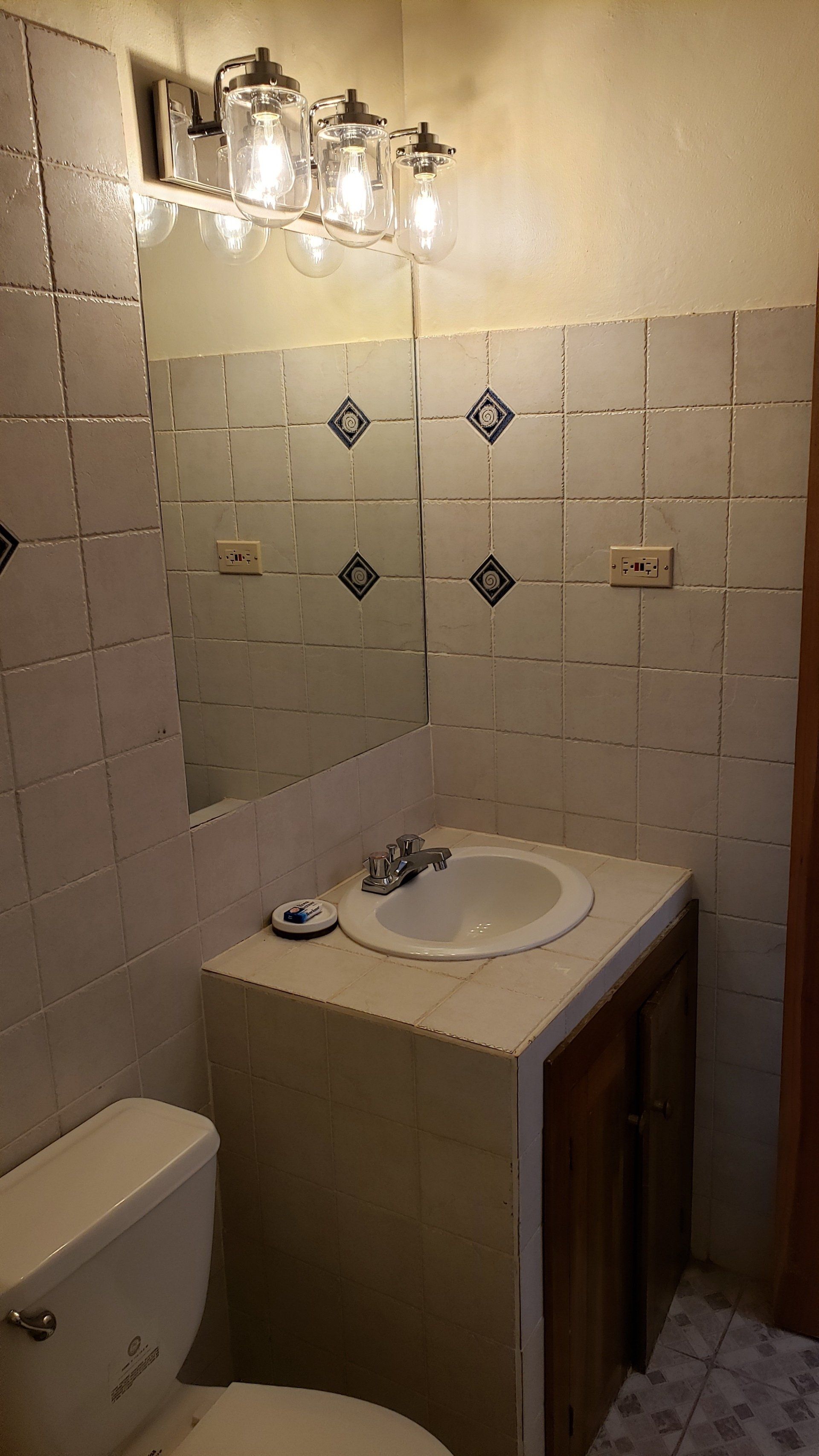 Bathroom with tiled walls, sink, mirror, toilet, and vanity cabinet. A light fixture is above the mirror.