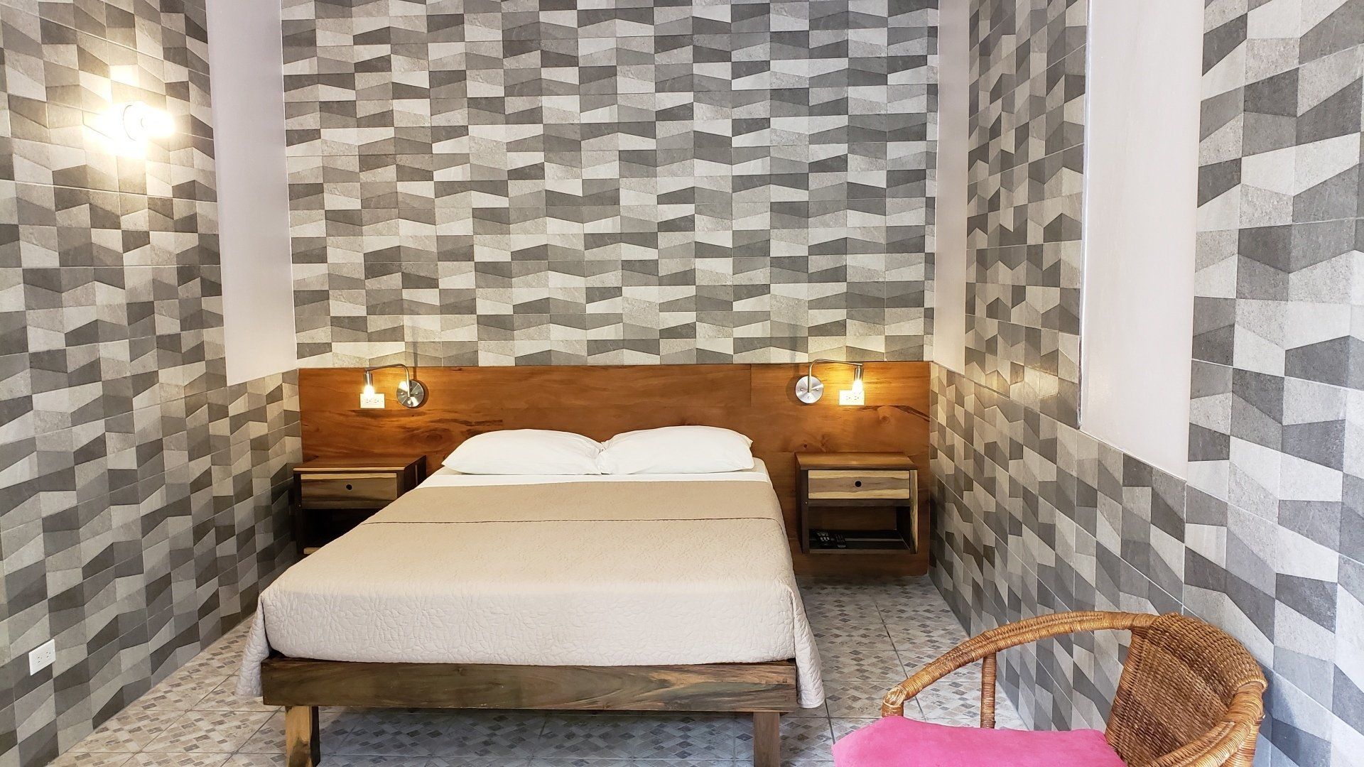 Bedroom with geometric patterned walls, wooden headboard, bed, bedside tables, wicker chair with pink cushion.