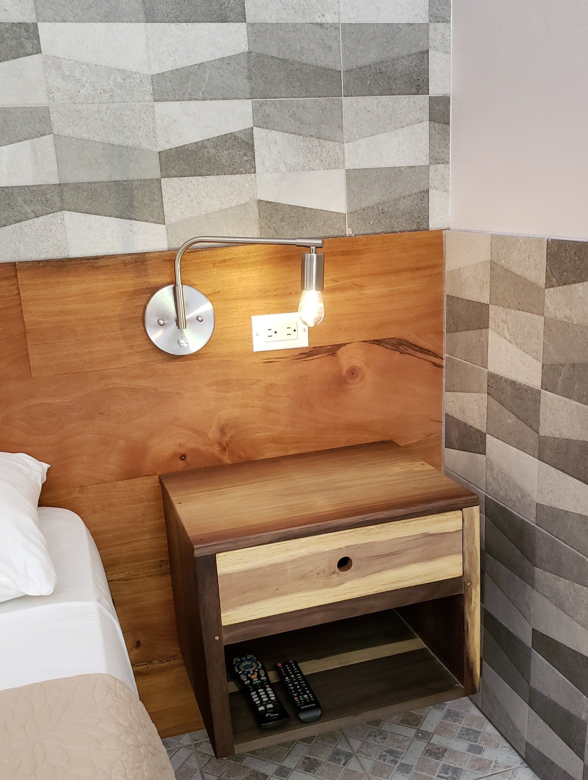 Wooden headboard and nightstand with a lamp and electrical outlet against patterned gray wall.
