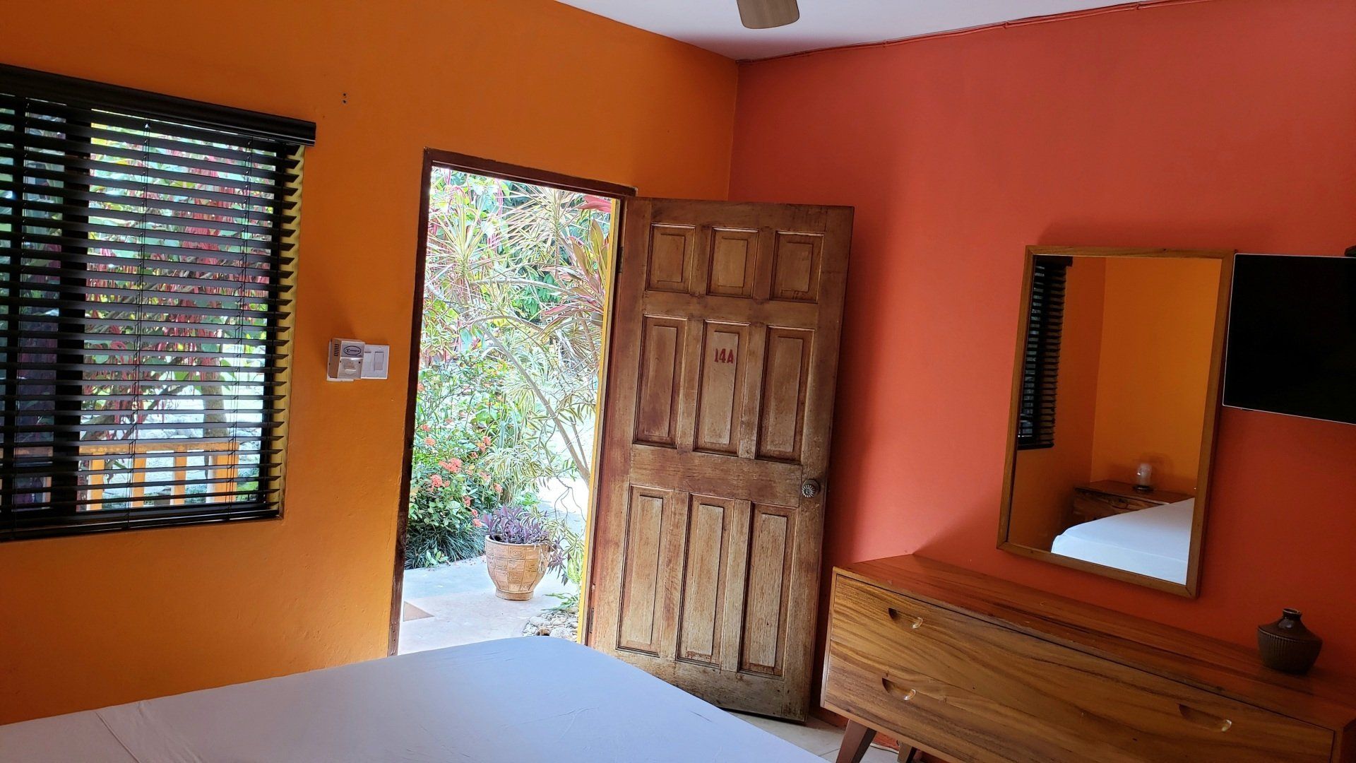 Orange bedroom with open wooden door to outside garden, wooden dresser with mirror and TV.