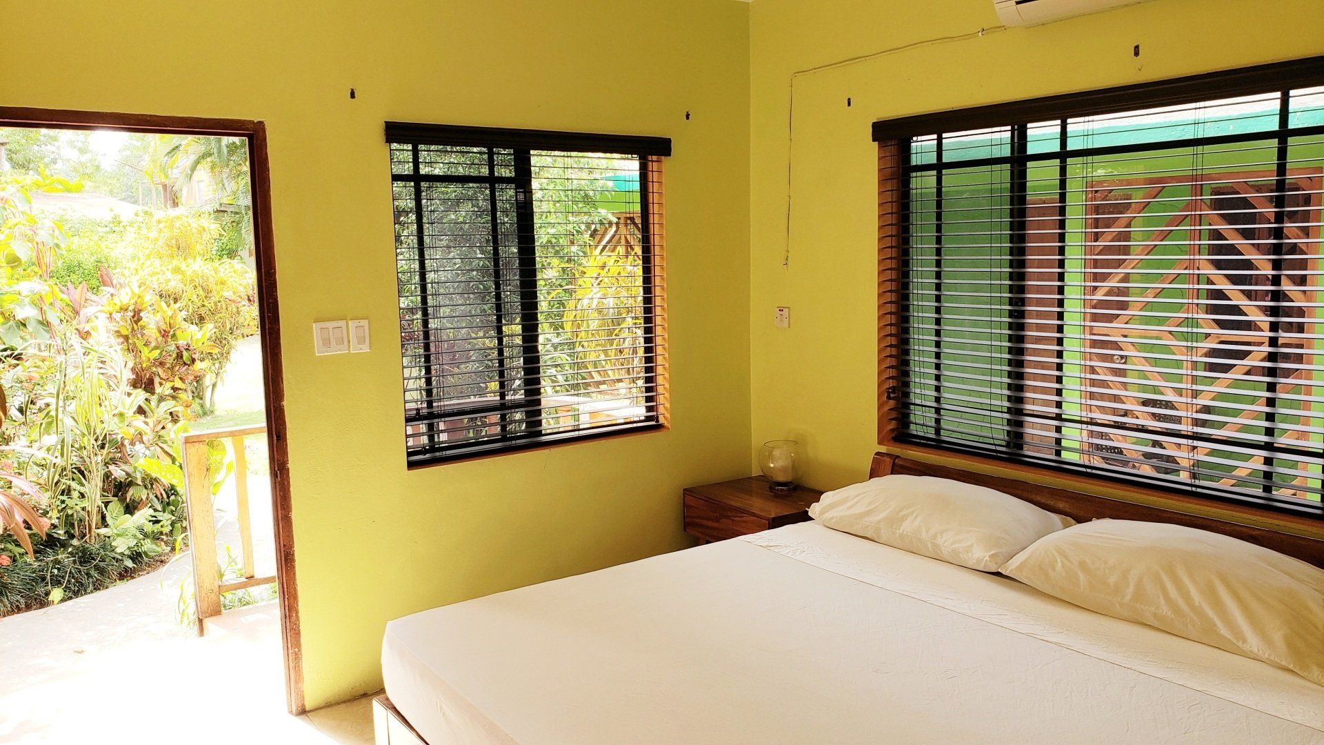 Bedroom with bed, open door, and windows overlooking greenery; walls are light yellow.
