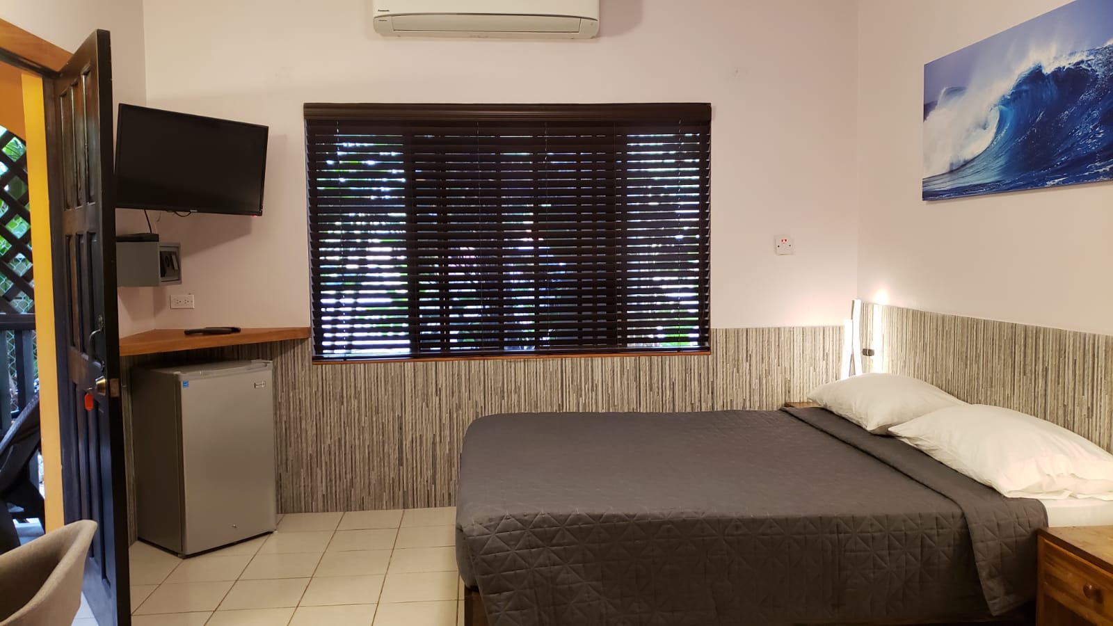 Hotel room with a bed, TV, mini-fridge, window, and wave art.