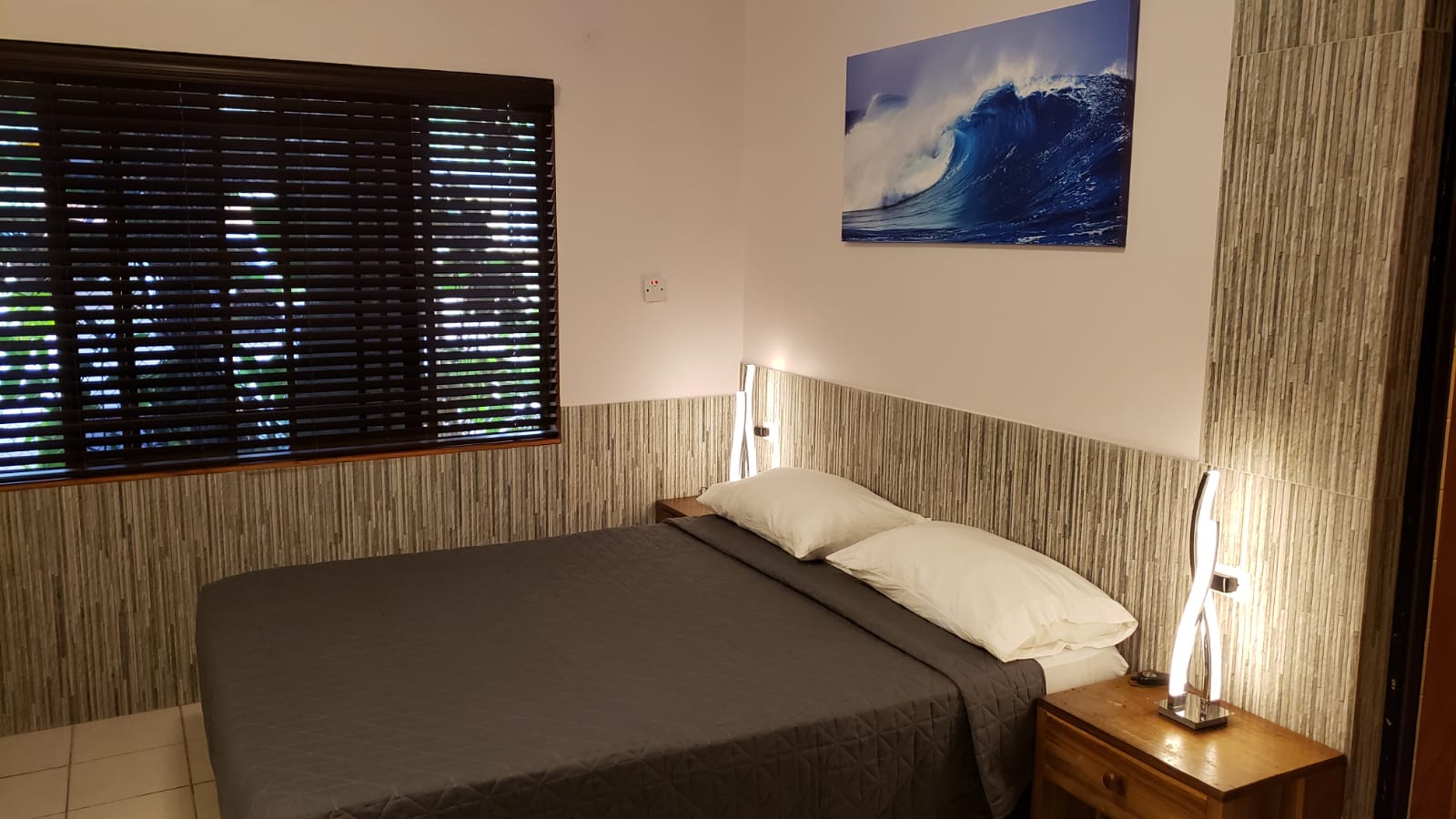 Bedroom with a bed covered in gray bedding, window, wood-grain walls, and a blue wave painting.