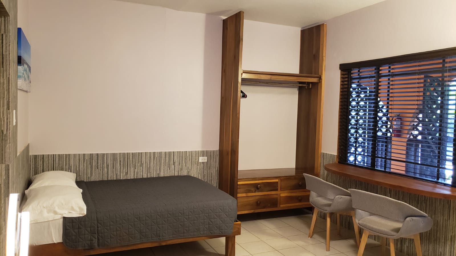 Bedroom with bed, closet, chairs, and window with blinds. Gray bedspread, wooden trim, white and gray walls.
