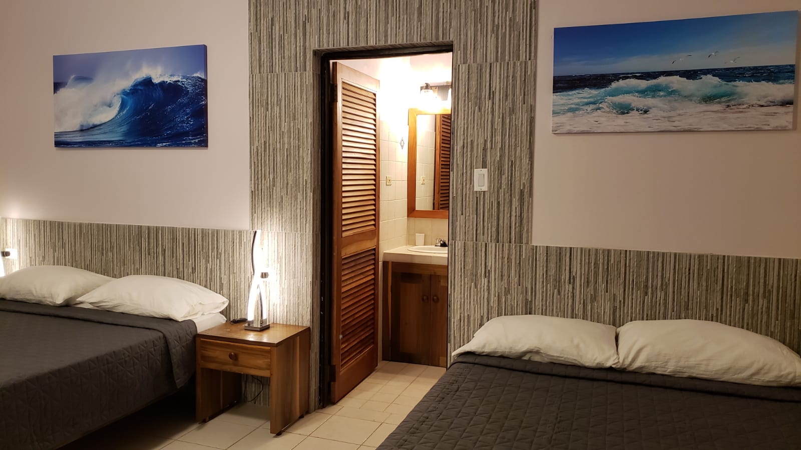 Two beds in a room with ocean wave art and a bathroom doorway. Gray bedding and light wood furniture.