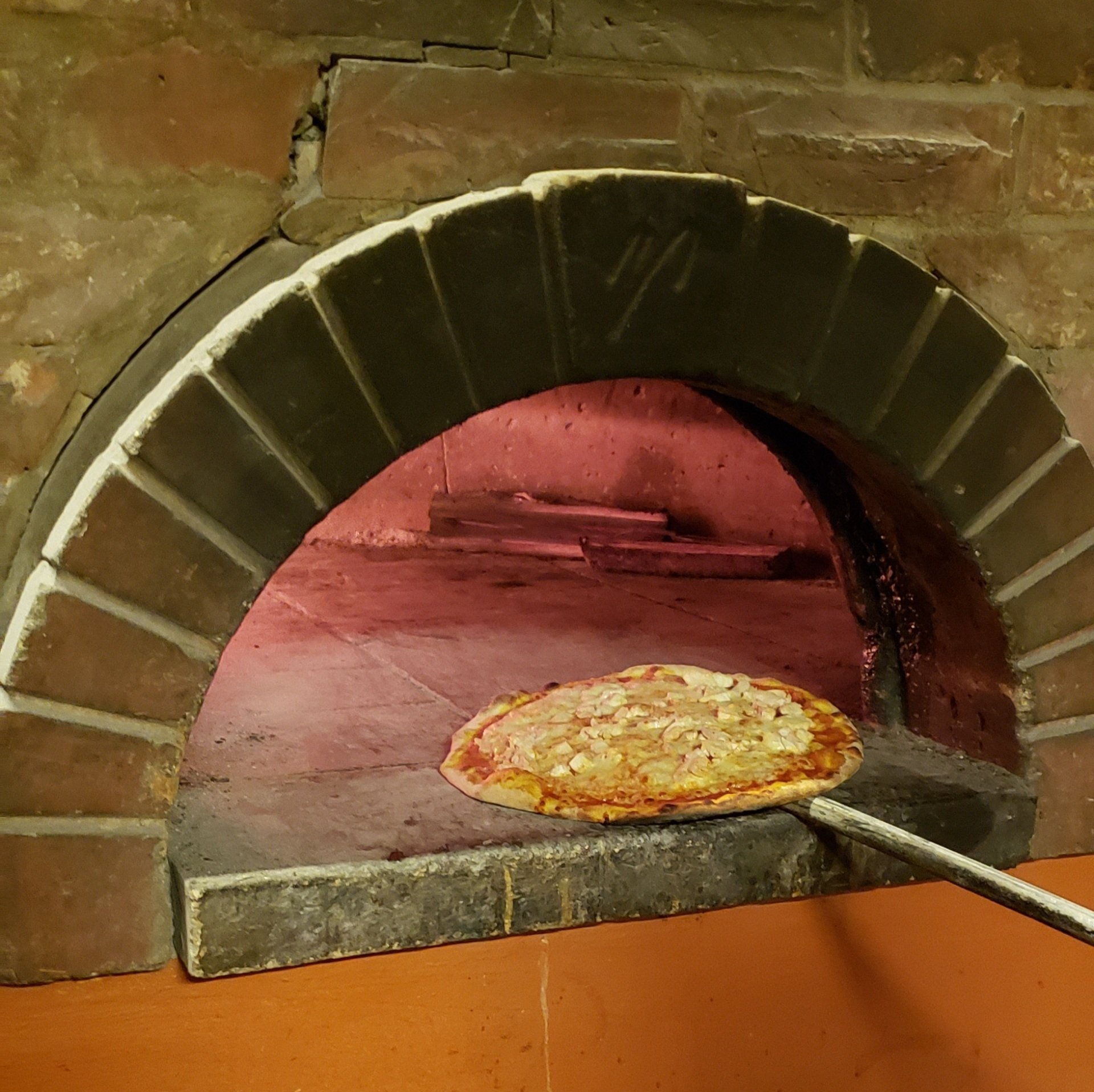 Pizza being removed from a brick oven with a long paddle; interior, reddish glow.