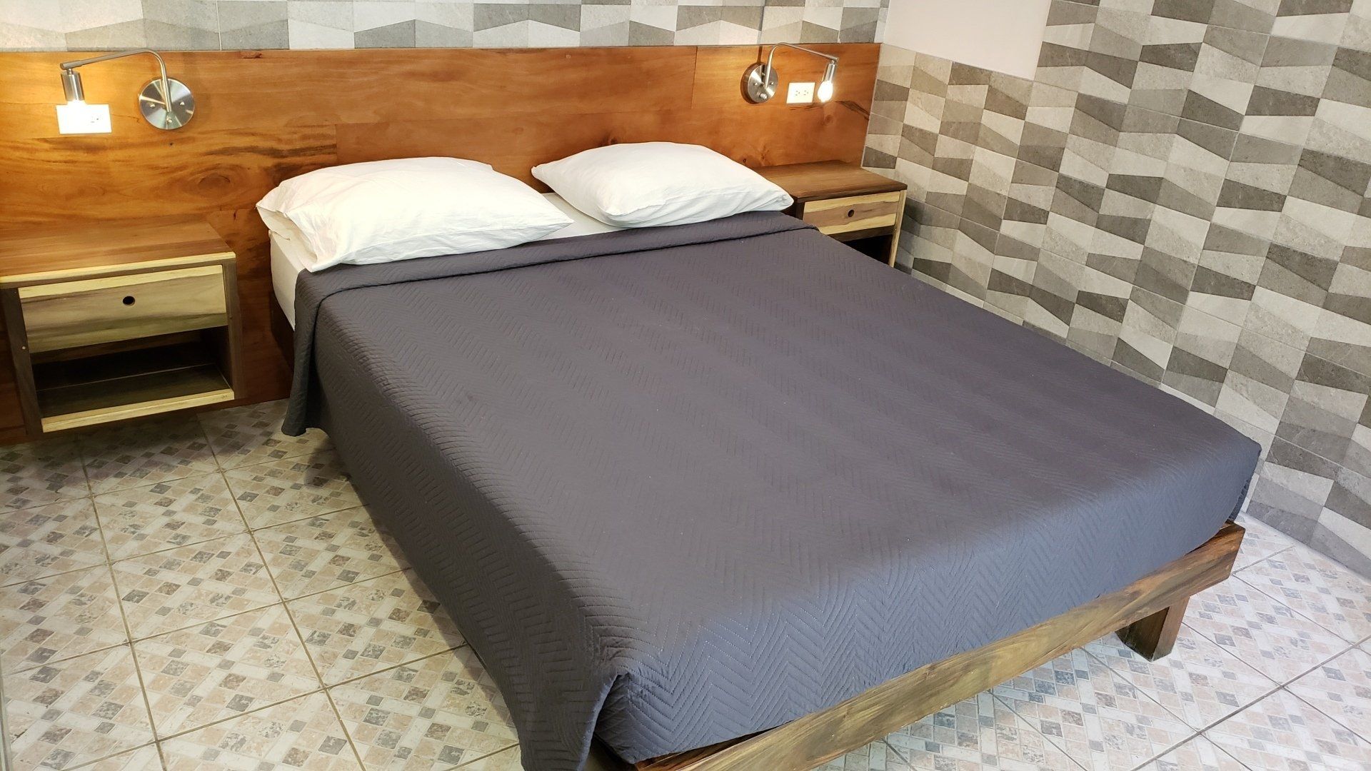 Bedroom with gray bedding, wooden headboard, two nightstands, and patterned wall tiles.