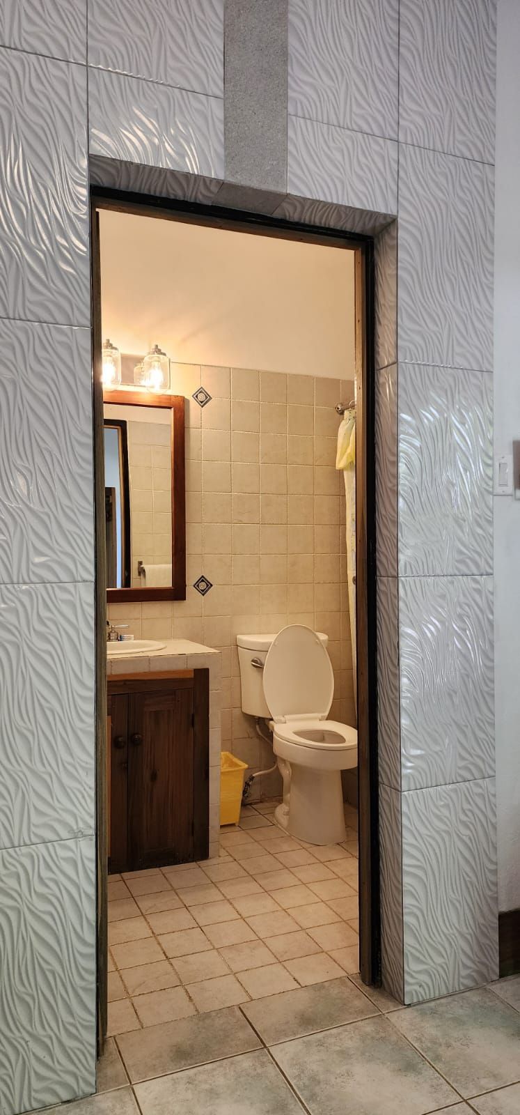 Bathroom doorway with tile walls. The room inside has a toilet, sink, mirror, and light fixtures.
