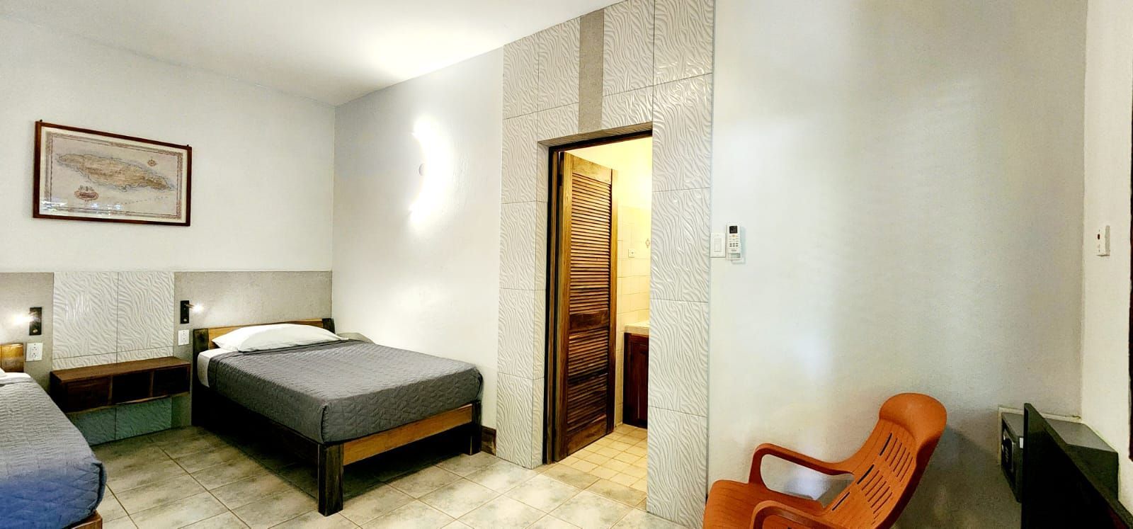 A sparsely furnished hotel room with two beds, an orange chair, and an open doorway.