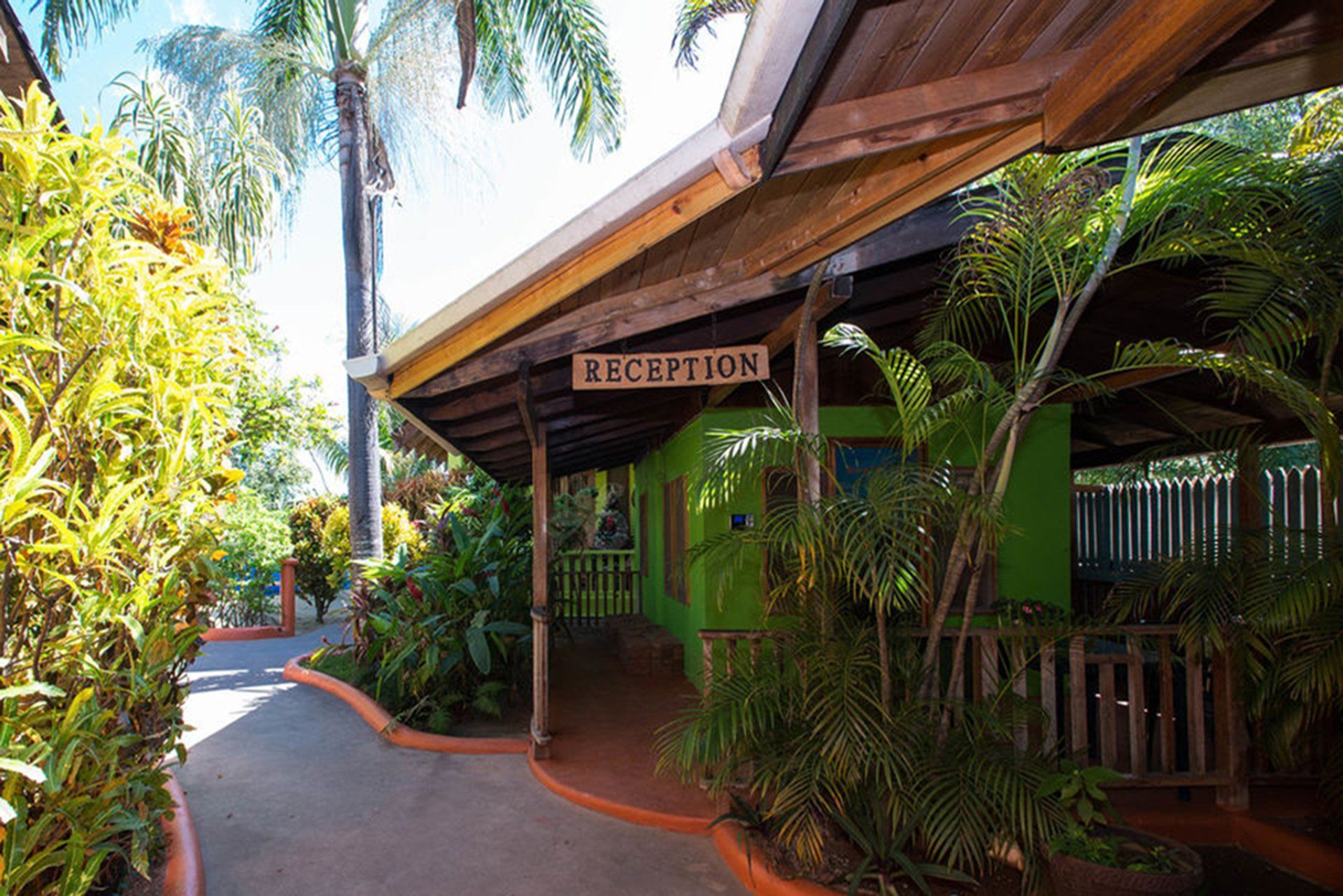 Reception area with green building, wooden beams, surrounded by lush greenery and a walkway.