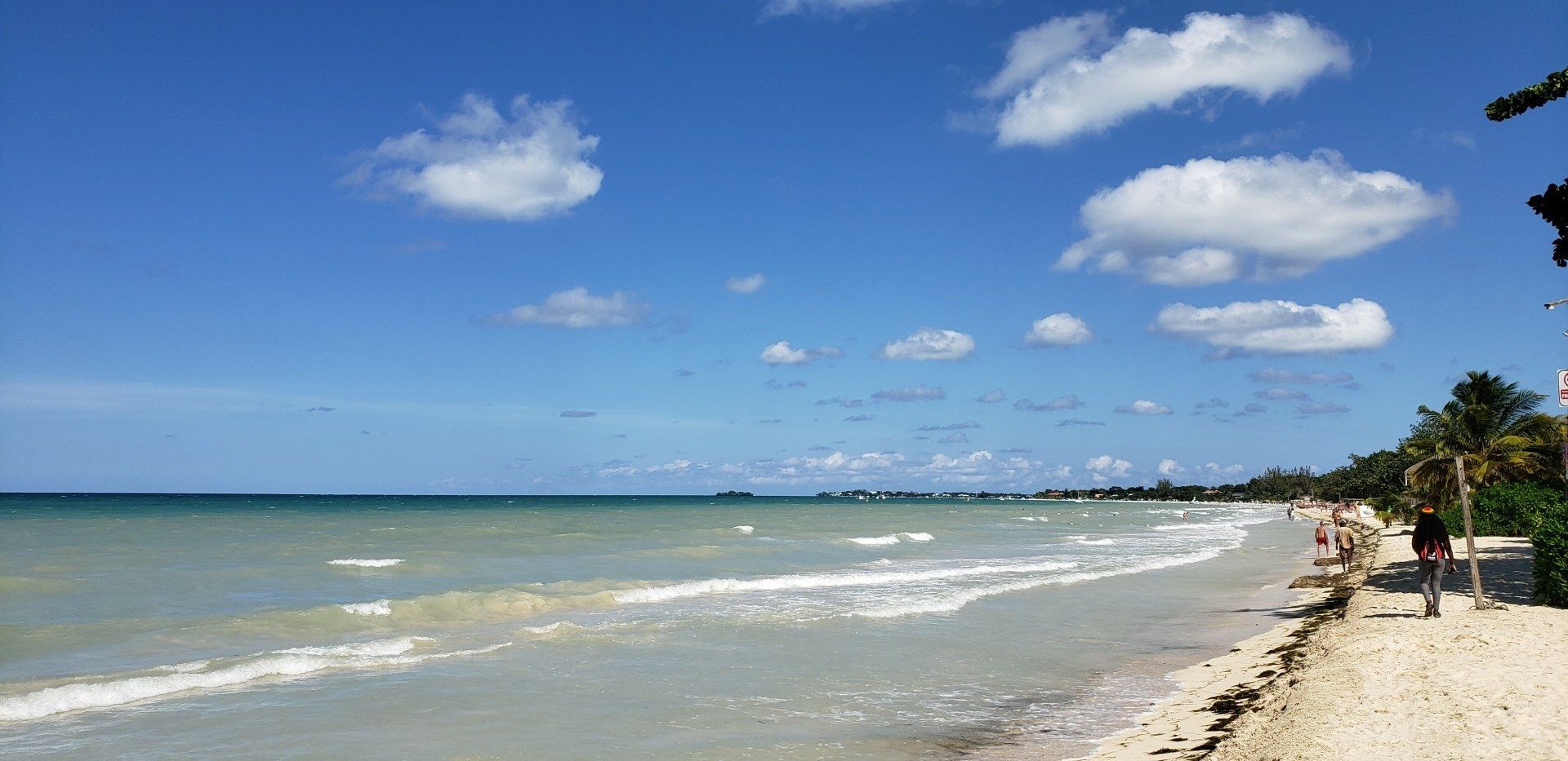 Beach with white sand, turquoise water, and fluffy clouds in a blue sky. People walk along the shore.