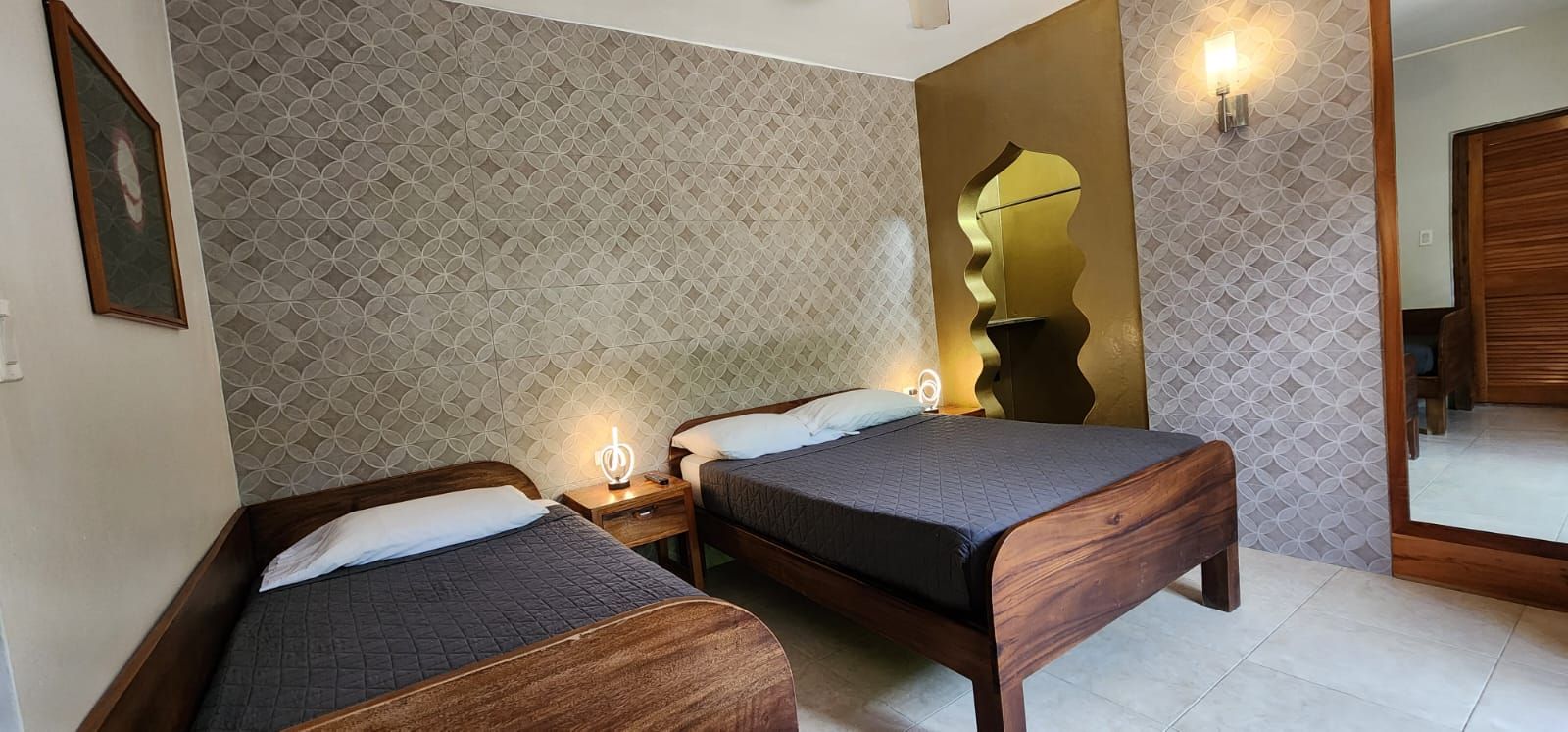 Bedroom with two beds, patterned wallpaper, a gold art piece, and a wooden framed mirror.