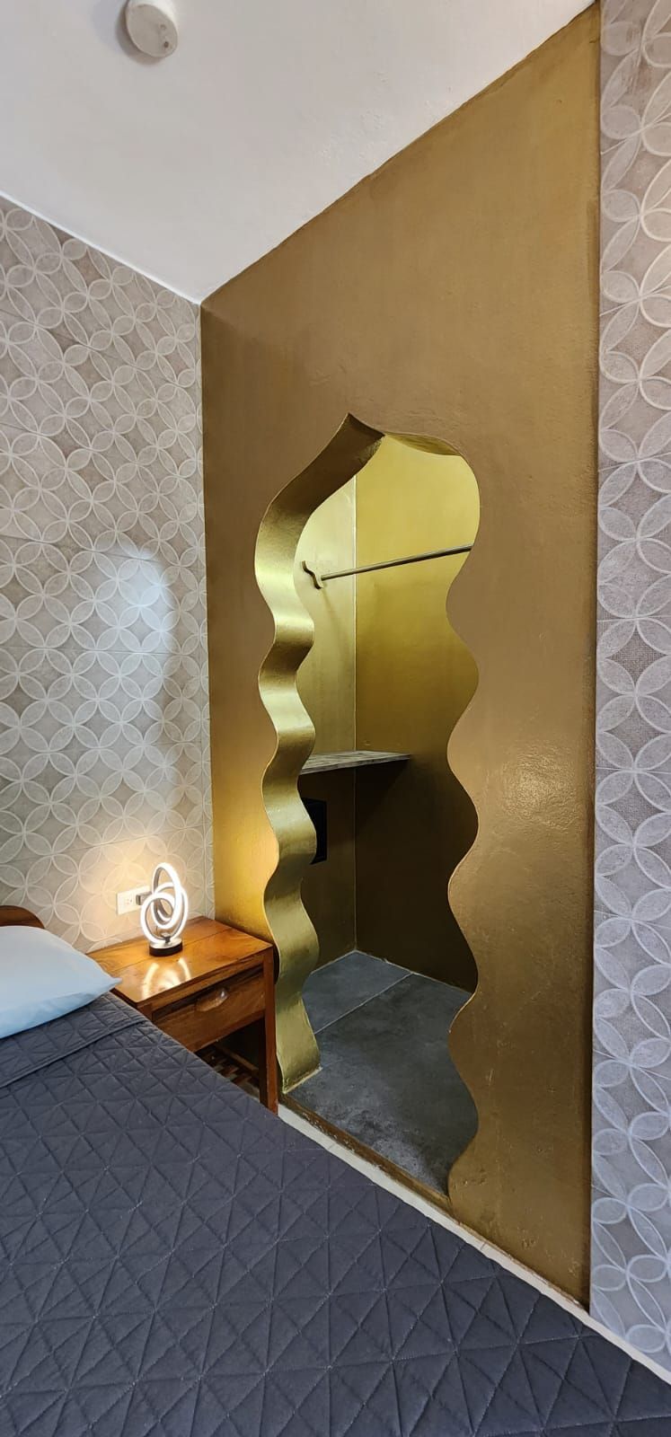 Bedroom with gold-painted wall, arched alcove, bedside table, bed with blue quilt, patterned wallpaper.