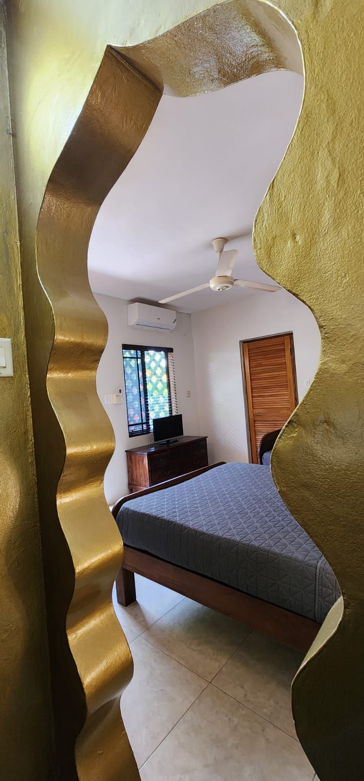 A wavy, gold-framed doorway reveals a bedroom. It includes a bed, tv, window, ceiling fan, and wooden door.