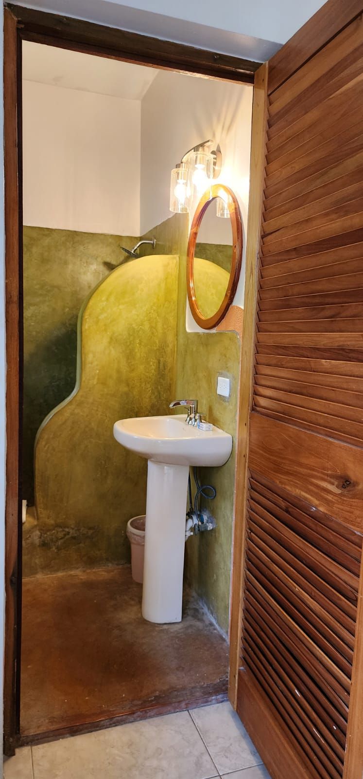 Bathroom interior: pedestal sink, oval mirror, green textured walls, wooden door.