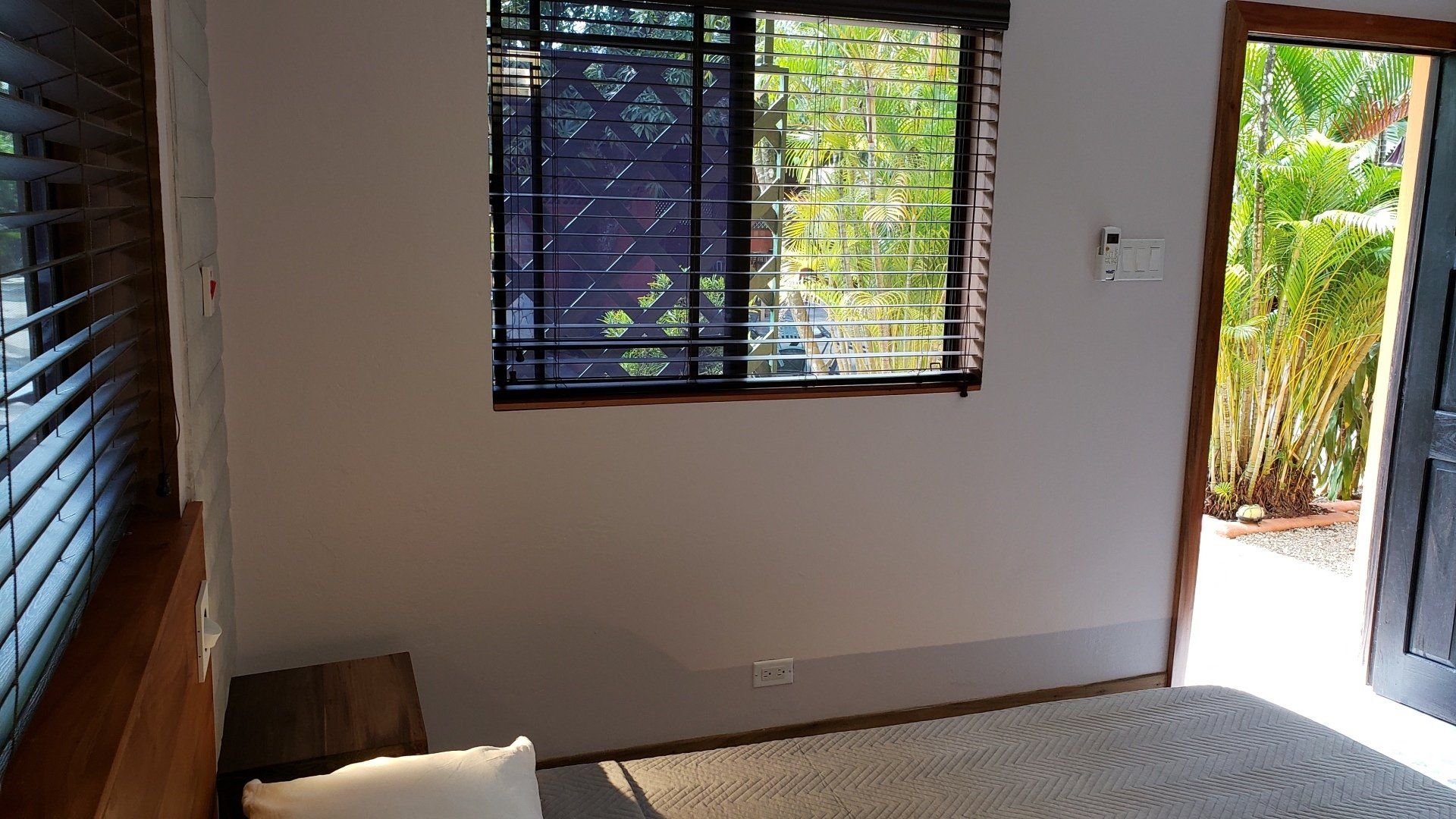 Bedroom with a bed, small window with blinds, and open door to greenery.
