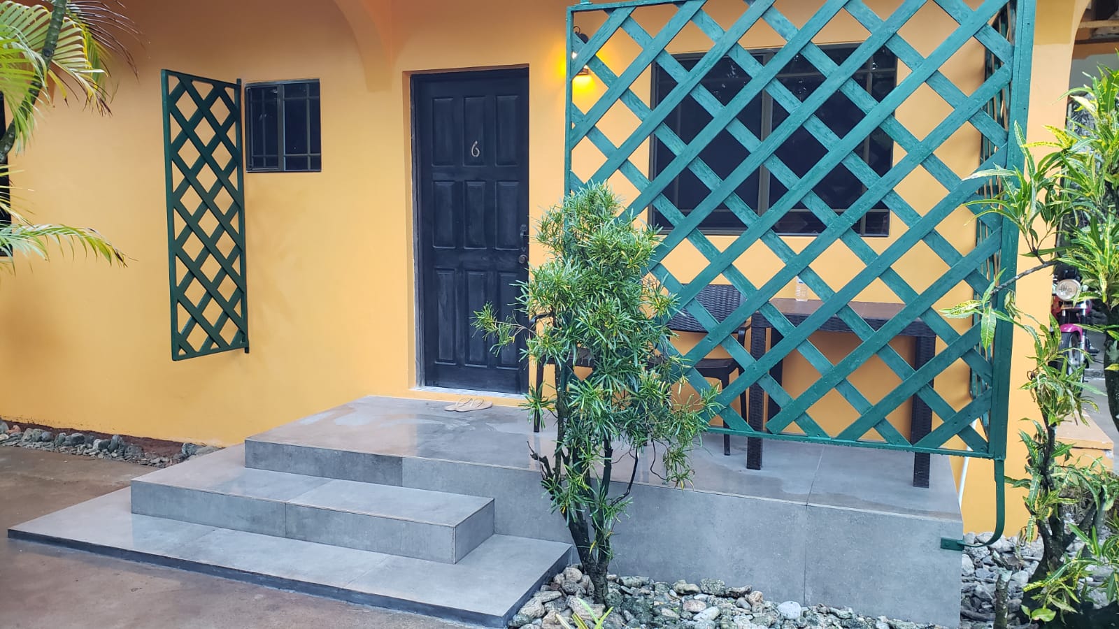 Yellow building with a black door, steps, and a green trellis. A table and chairs are behind the trellis.