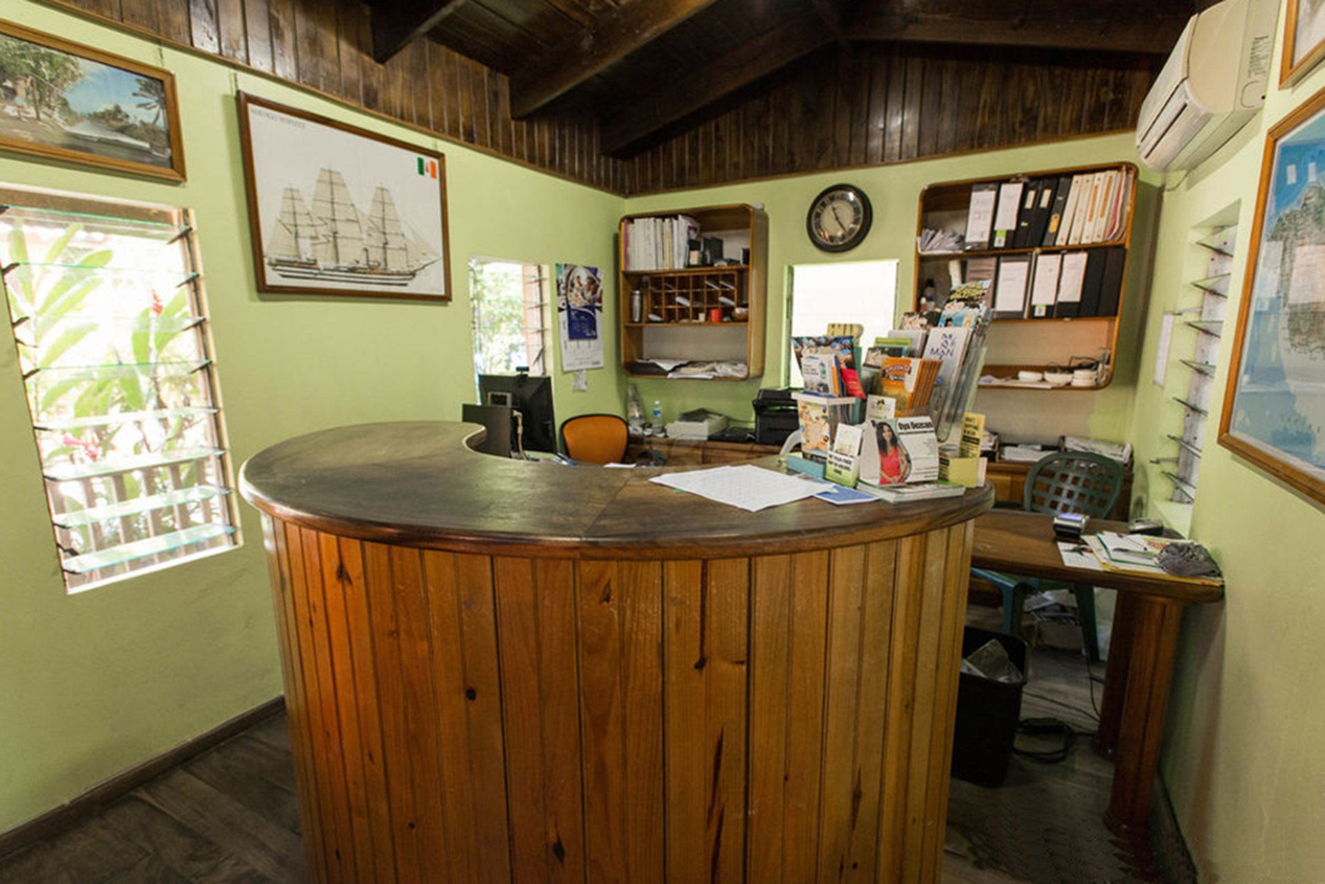 Wooden reception desk in a small room with light green walls. Artwork, papers, and office supplies are visible.