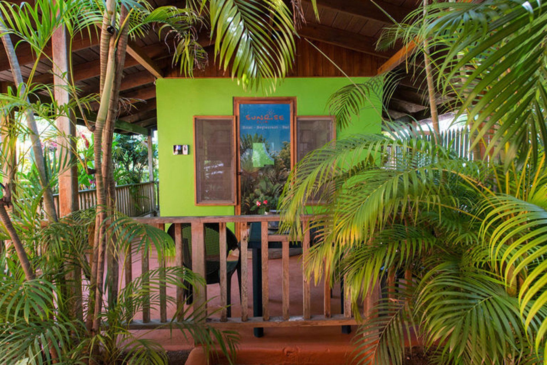 Green painted building with a sign framed by brown wood, and surrounded by tropical plants.
