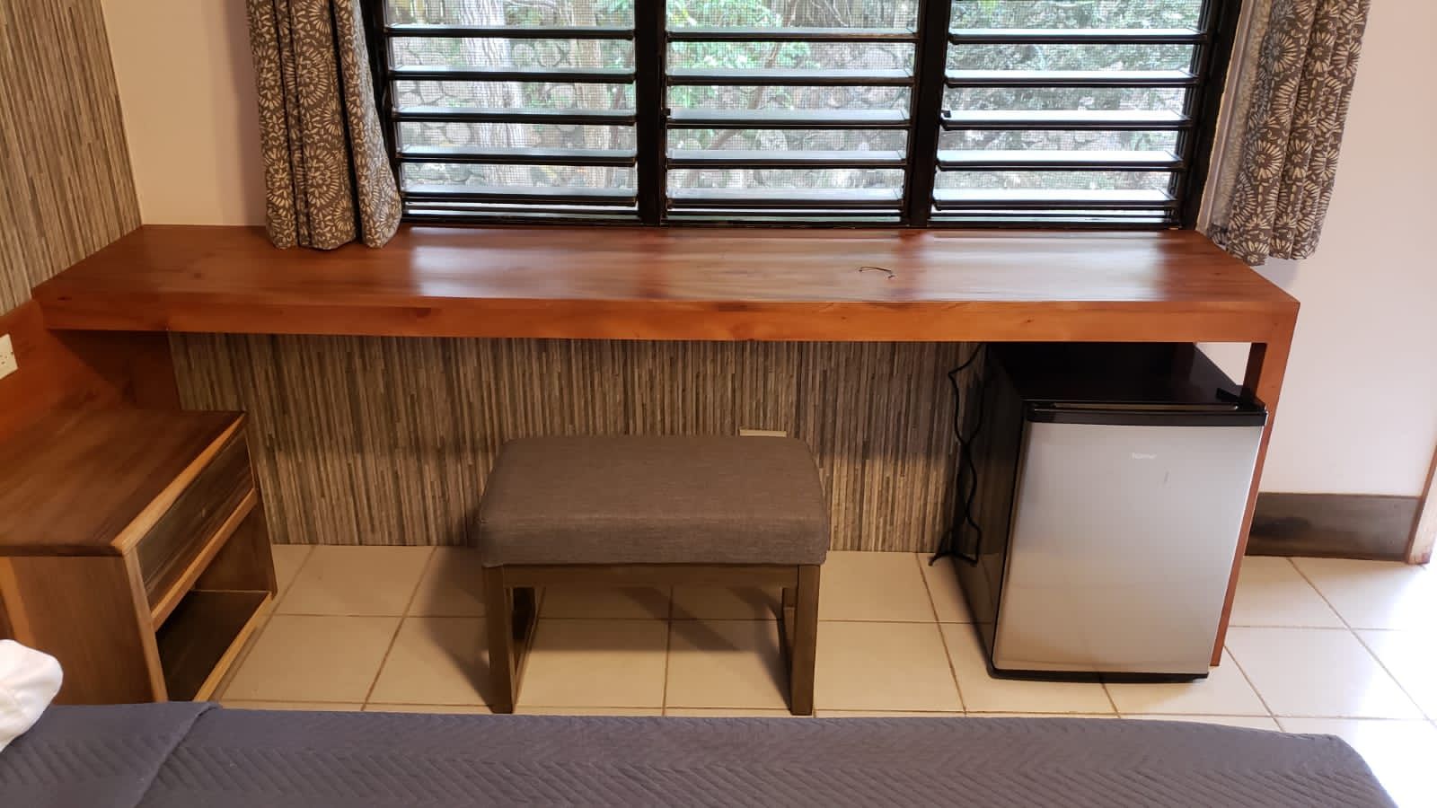 Wooden desk with a stool, mini-fridge, and window, with a nightstand to the side.