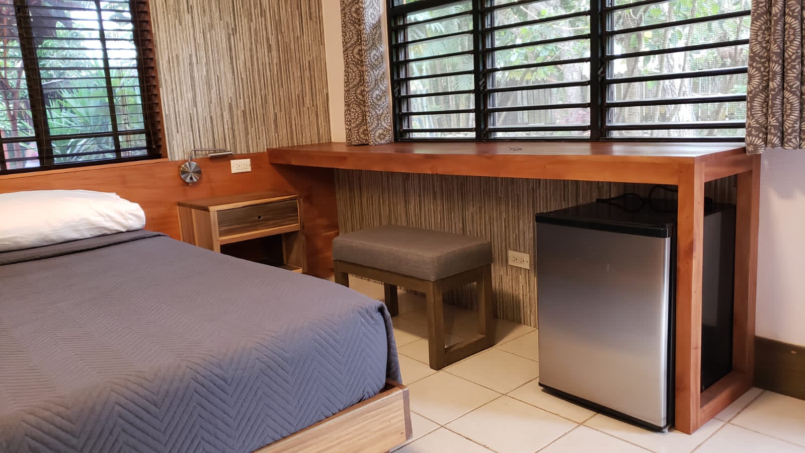 Bedroom with bed, desk, stool, and small refrigerator. Window with shutters.